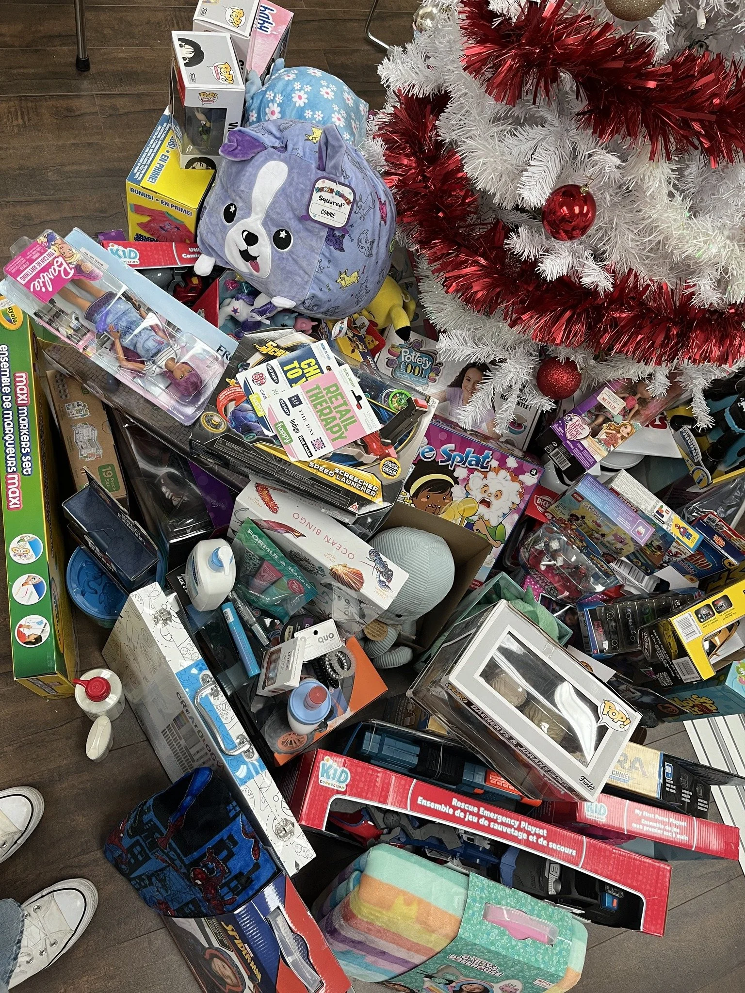 A collection of toys and holiday decorations including a white Christmas tree with red and white garland, red ornaments, and wrapped gifts. Items on the floor include dolls, games, and plush toys.
