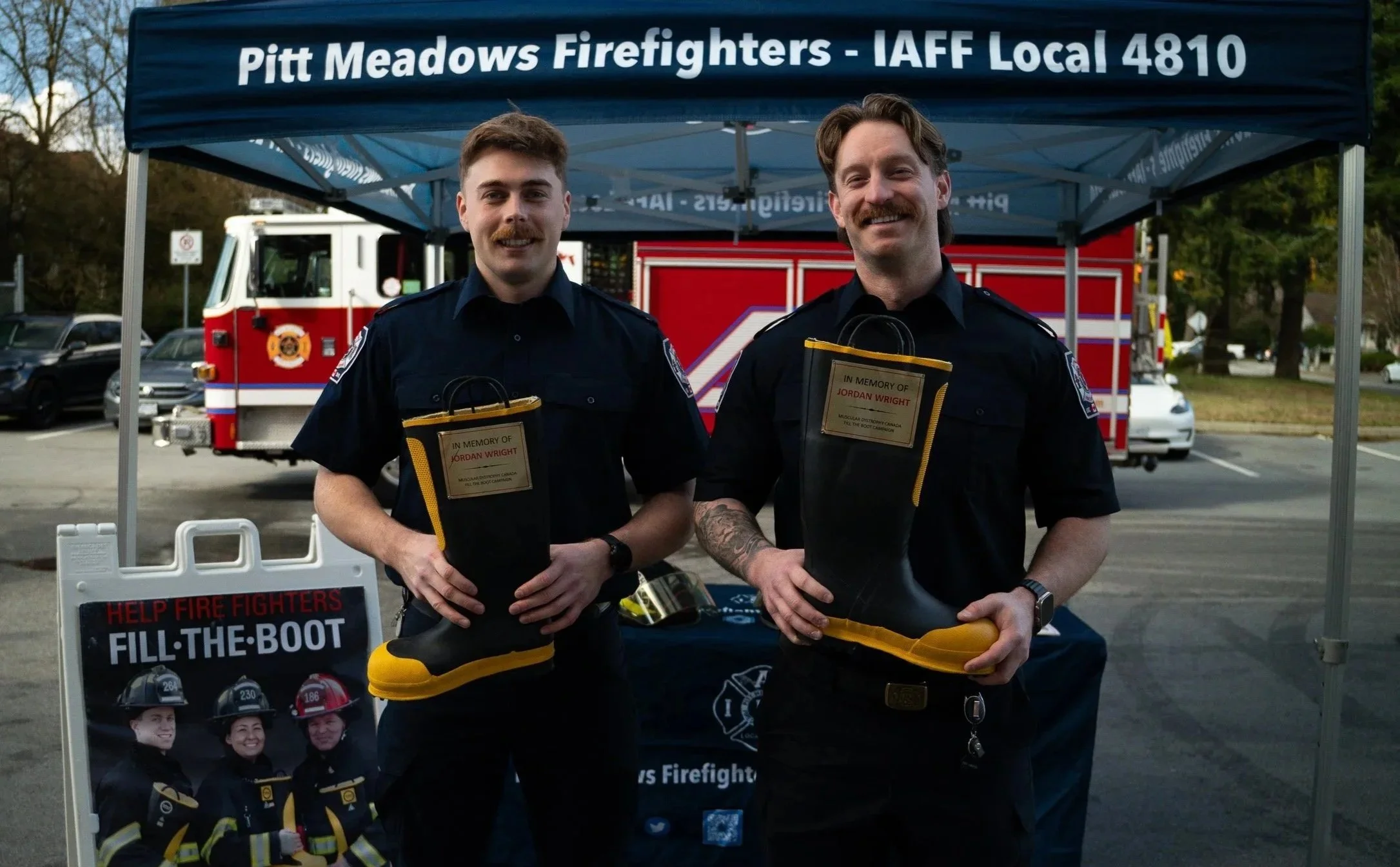 Two male firefighters standing under a blue canopy with Pitt Meadows Firefighters and IAFF Local 4810 written on it, smiling and holding firefighter boots, with a fire truck in the background.