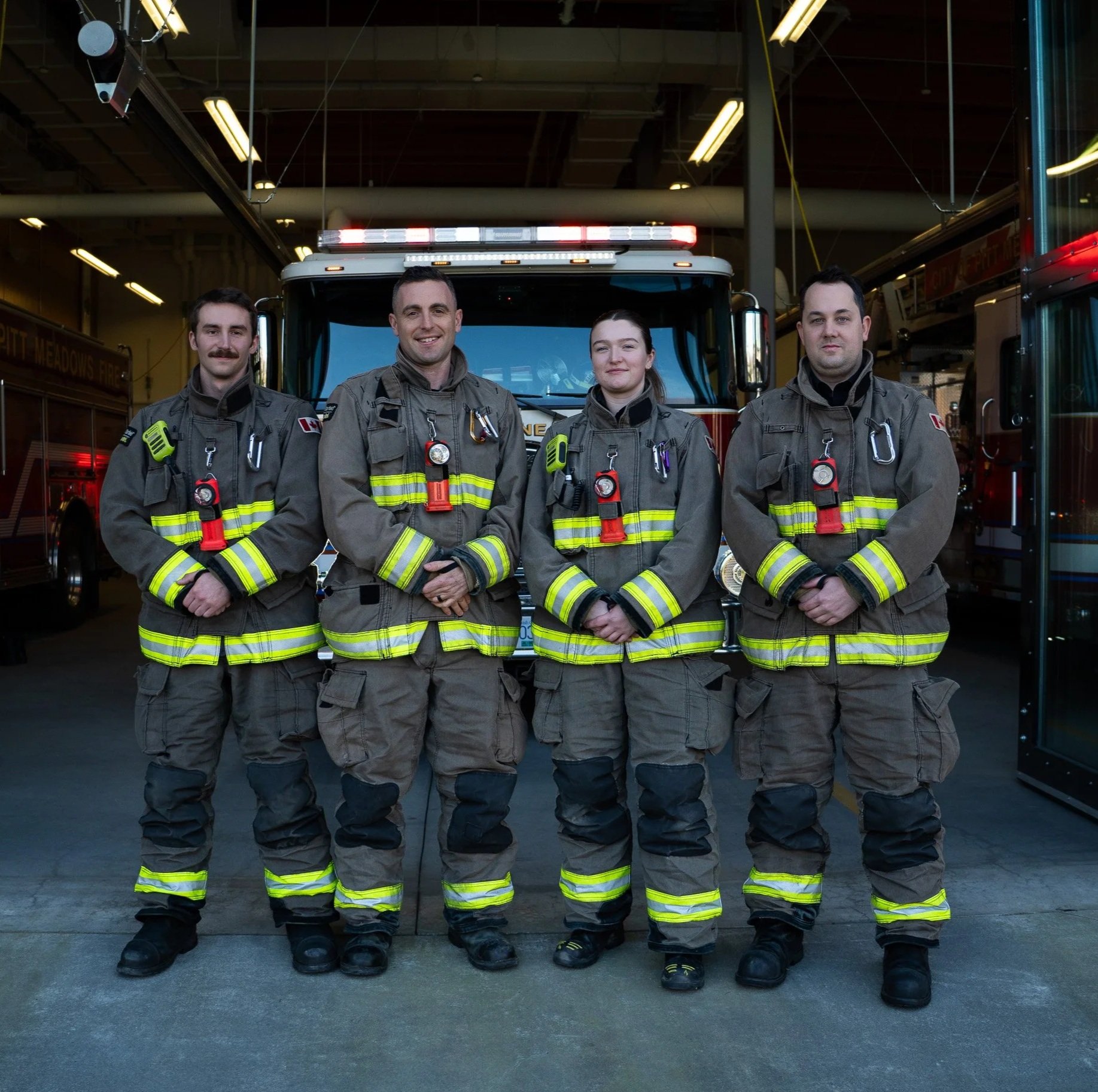 Four firefighters in uniform standing in front of a fire truck in a fire station.