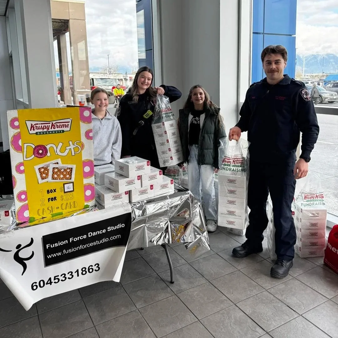 Group of four people standing inside a building near a table with boxes of Krispy Kreme doughnuts, participating in a fundraiser.
