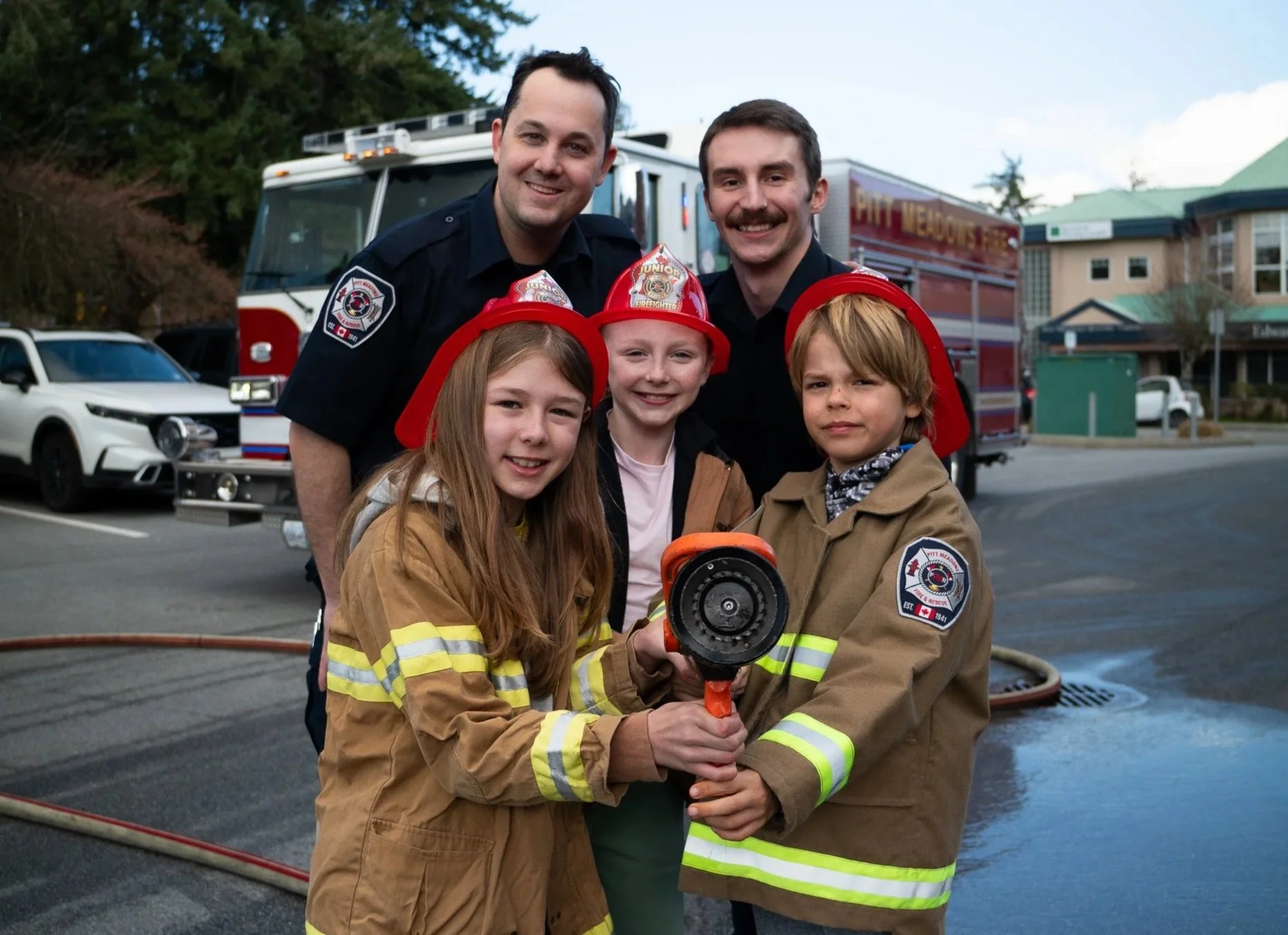 Two firefighters and three children smiling at the camera, holding a fire hose nozzle. The children are wearing red firefighter helmets, and the firefighters are dressed in firefighter uniforms, standing in front of fire trucks with houses in the background.