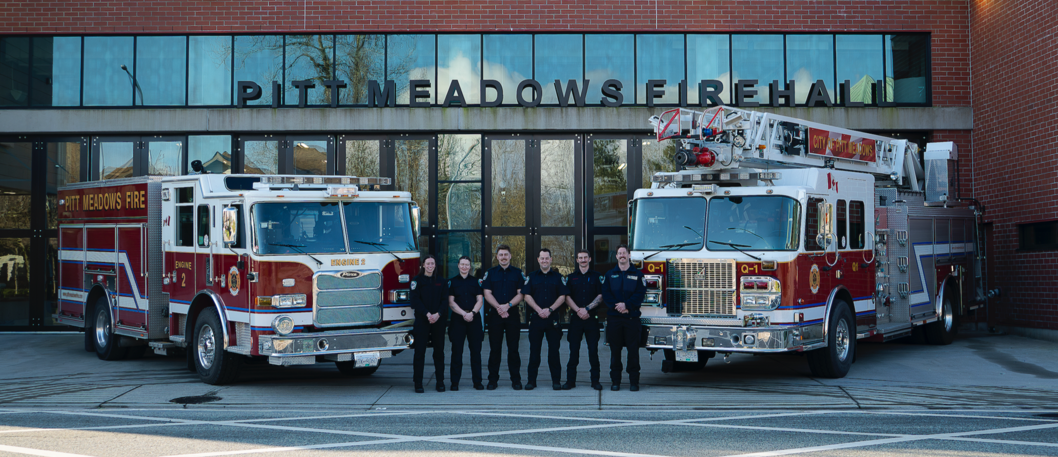 Six firefighters standing in front of fire trucks outside Pitt Meadows Firehall.