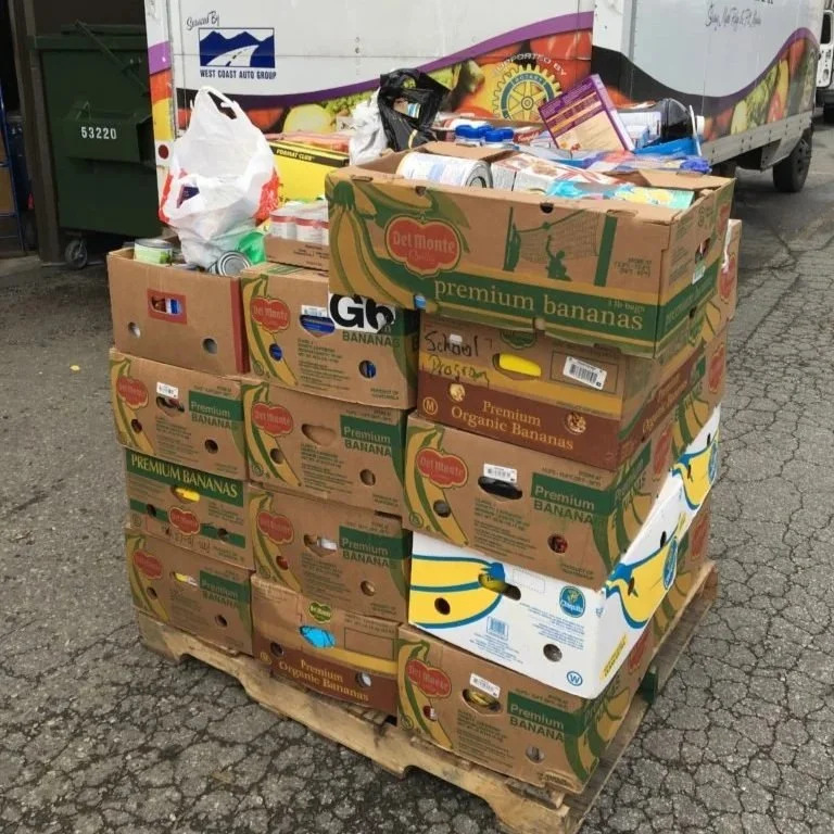A pallet stacked with boxes of Del Monte organic bananas and other produce, located outdoors next to a cargo truck and a green dumpster.