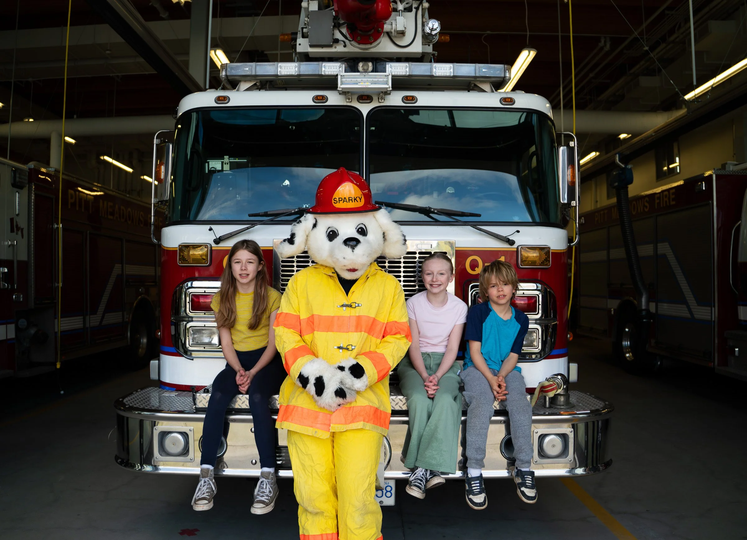 A person dressed as Sparky the firefighter dog mascot with a Dalmatian costume and firefighter uniform standing in front of a fire truck with children sitting on the front. The fire truck is inside a fire station bay.