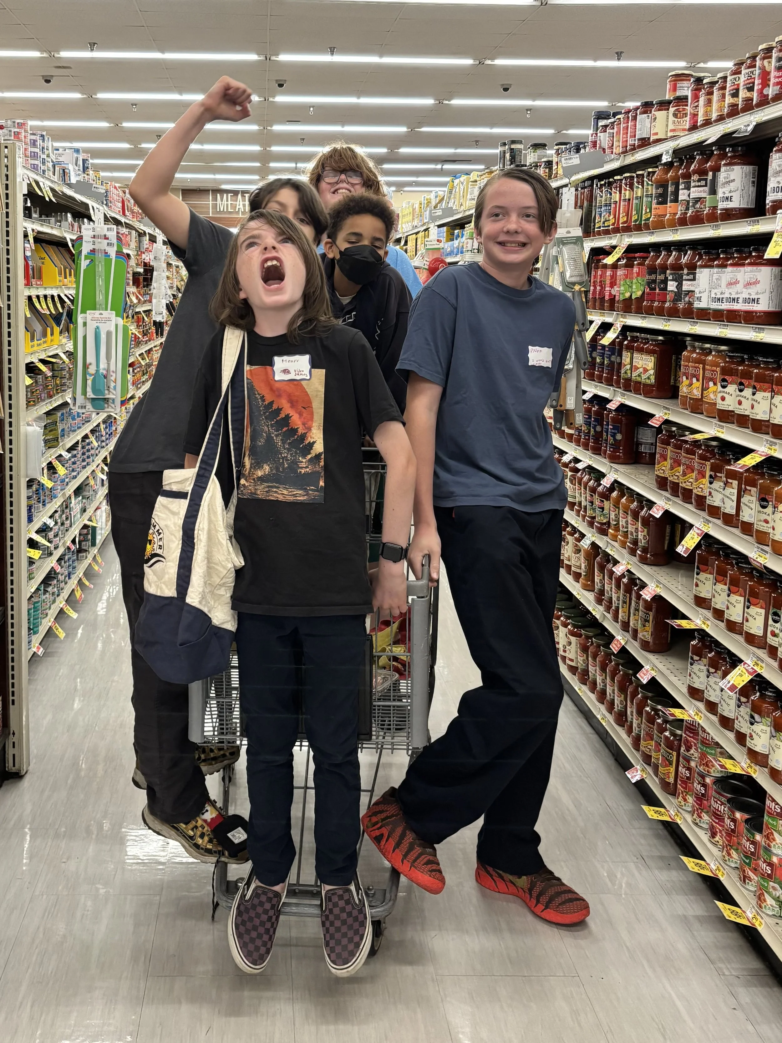 Group of five children standing and sitting in a grocery store aisle, posing with playful expressions and gestures, surrounded by shelves stocked with jars of pasta sauce.