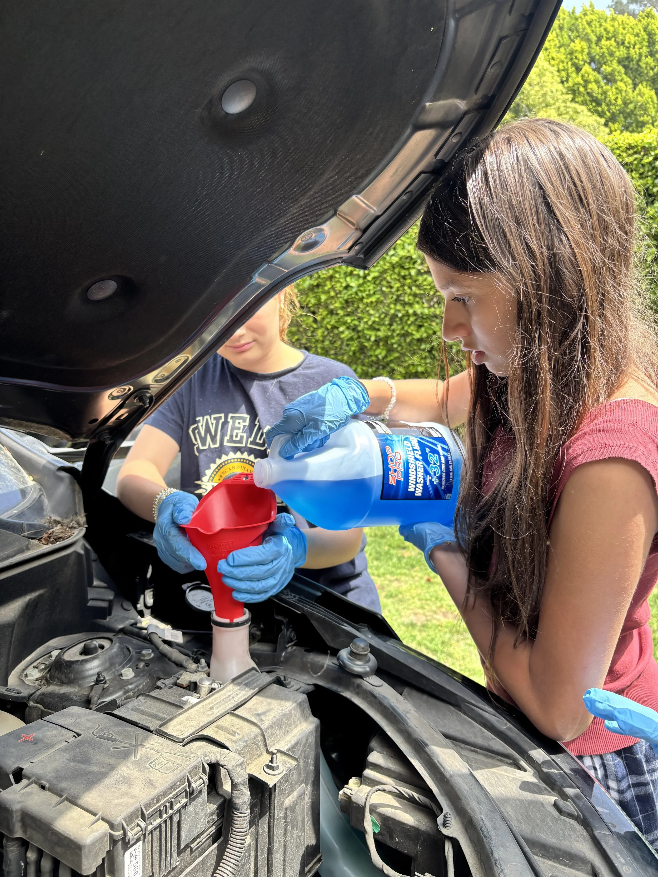 Two young girls are working on fixing a car engine. One girl is pouring a blue liquid into the engine with a funnel, while the other girl is holding the funnel steady. Both girls are wearing blue gloves. The car's hood is open, revealing the engine components, and there are trees and greenery in the background.