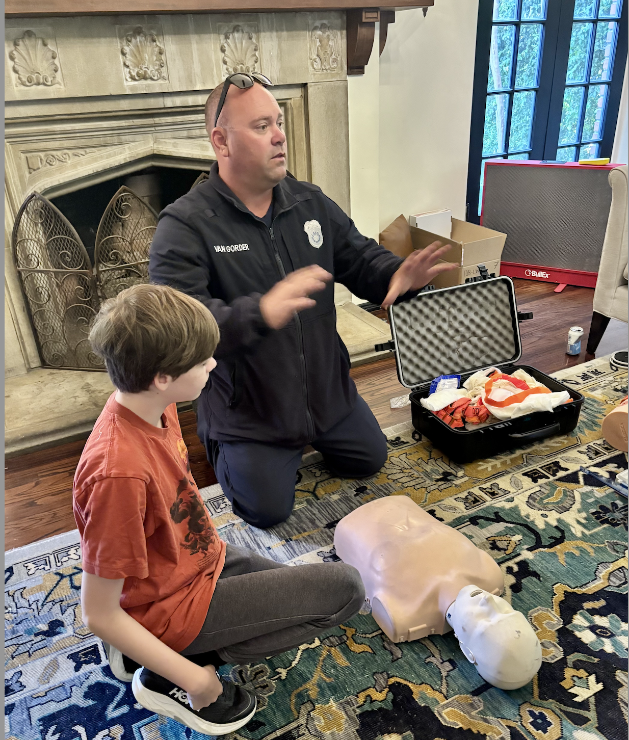 A man in a black jacket with a police badge explaining CPR techniques to a young boy in a red shirt. They are kneeling on a patterned rug in front of a fireplace, with a CPR mannequin and a medical equipment case open nearby.