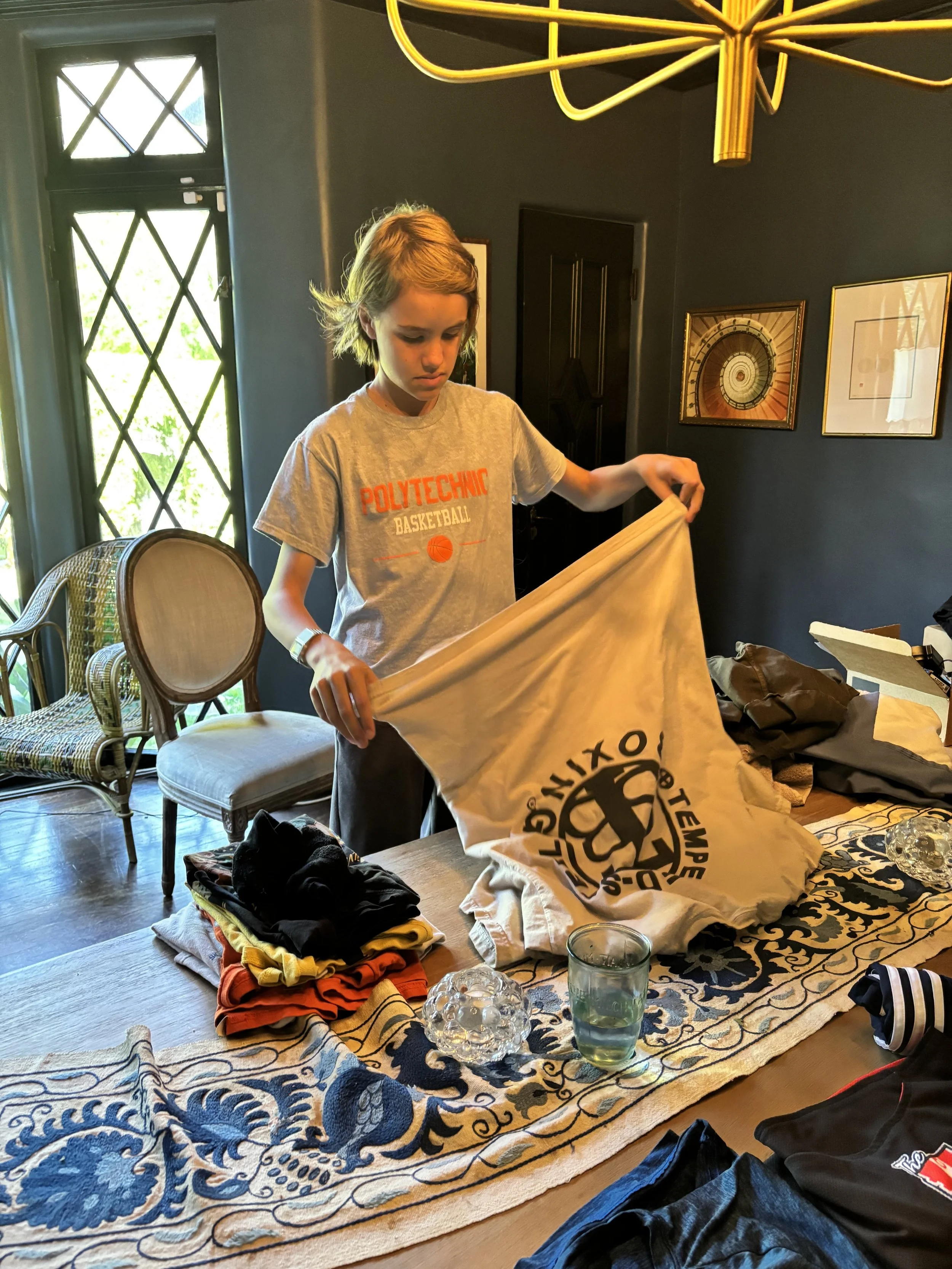 A boy with shoulder-length blonde hair wearing a gray T-shirt with 'Polytechnic Basketball' printed on it, standing at a table with clothes, a bag, and a plastic bag. He is holding a large yellow bag with a black logo. In the background, there are two chairs with woven seats, a window with diamond-shaped panes, and framed artwork on dark-colored walls.