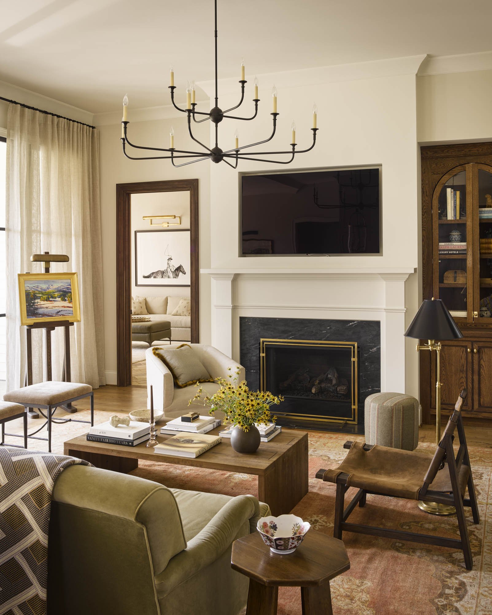 Interior of a living room with a fireplace, wall-mounted TV, chandelier, and furniture including armchairs, sofa, and coffee table with books and a vase of flowers.