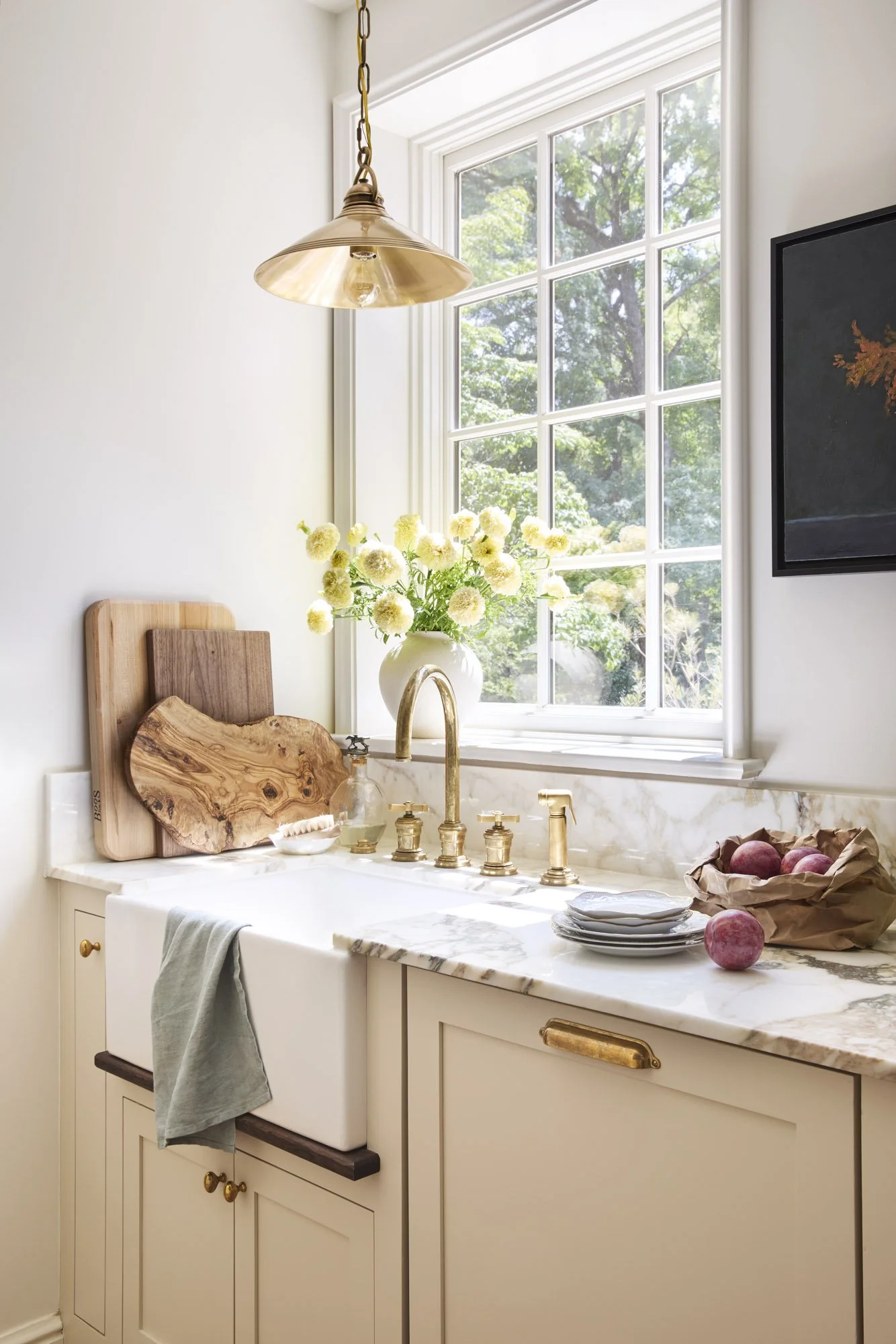 A kitchen with a large window displaying green trees outside. A white farmhouse sink with a brass faucet is set into a marble countertop. Decor items include a vase with yellow flowers, cutting boards, and a bowl of peaches, with a hanging gold light