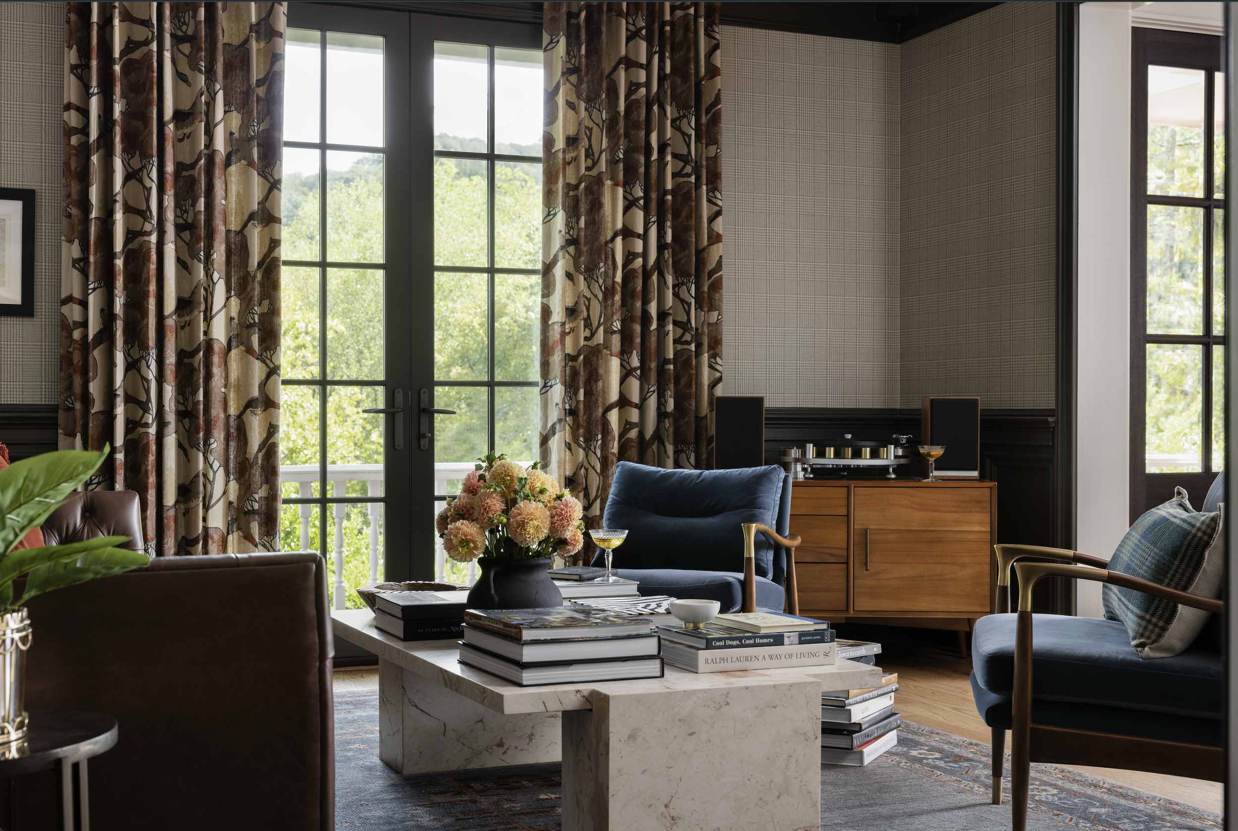 Living room with large windows and glass double doors, floral curtains, blue and brown chairs, a coffee table with books and flowers, and a wooden sideboard. Reflects a cozy, elegant space with greenery outside.