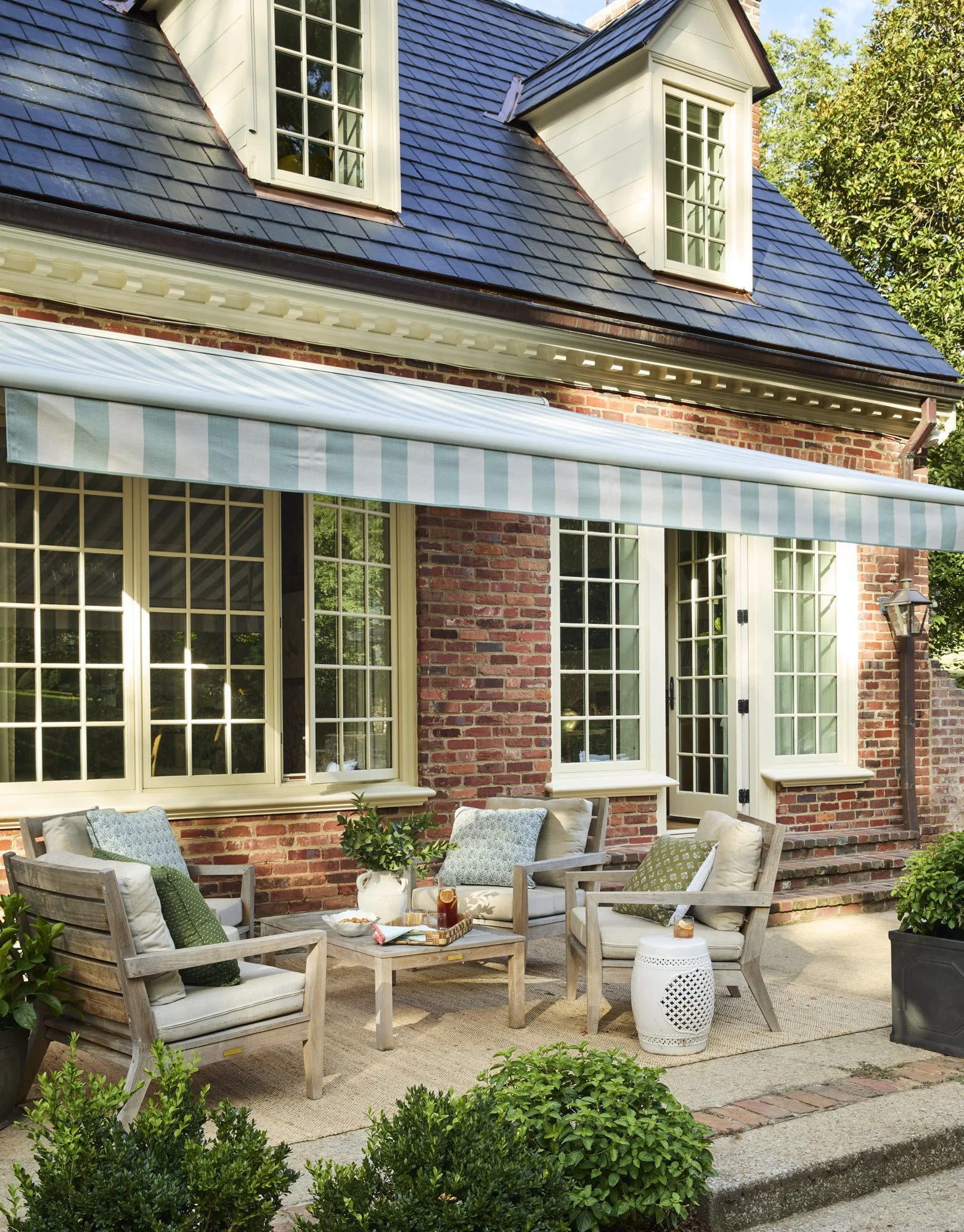 A cozy outdoor patio with wooden chairs and a table set on a rug. Potted plants surround the seating area in front of a brick house with large windows, a striped retractable awning, and French doors. Green trees are visible in the background.