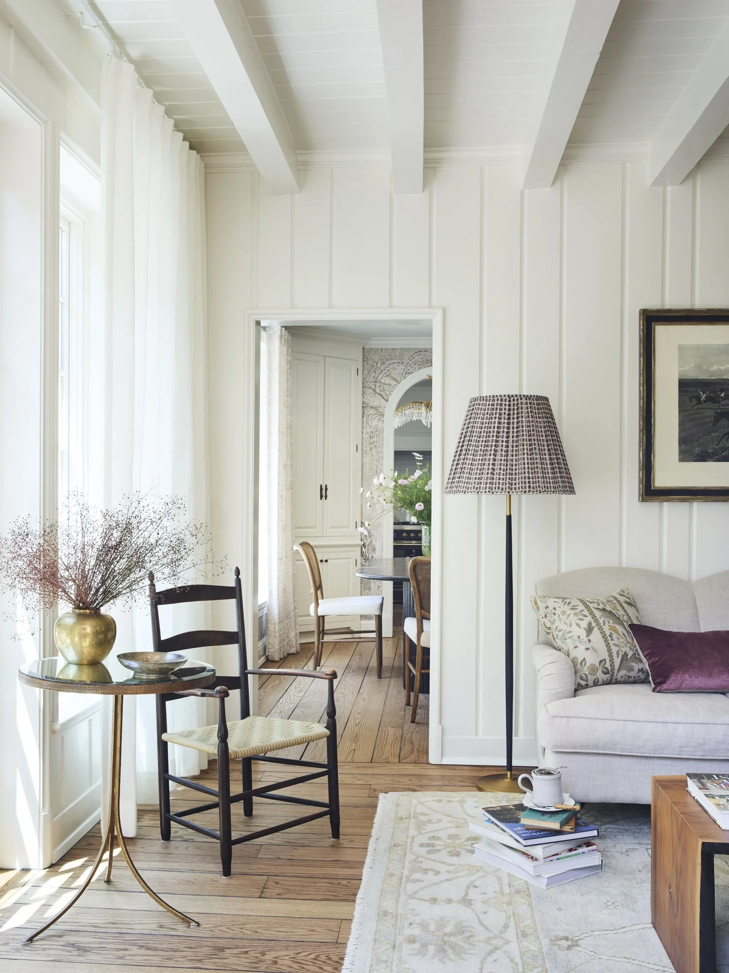 Living room with white walls, large windows with curtains, and hardwood floors. There is a small round table with a gold vase and a black chair. A beige sofa with pillows and a floor lamp are also visible. Through a doorway, a dining area with a tabl