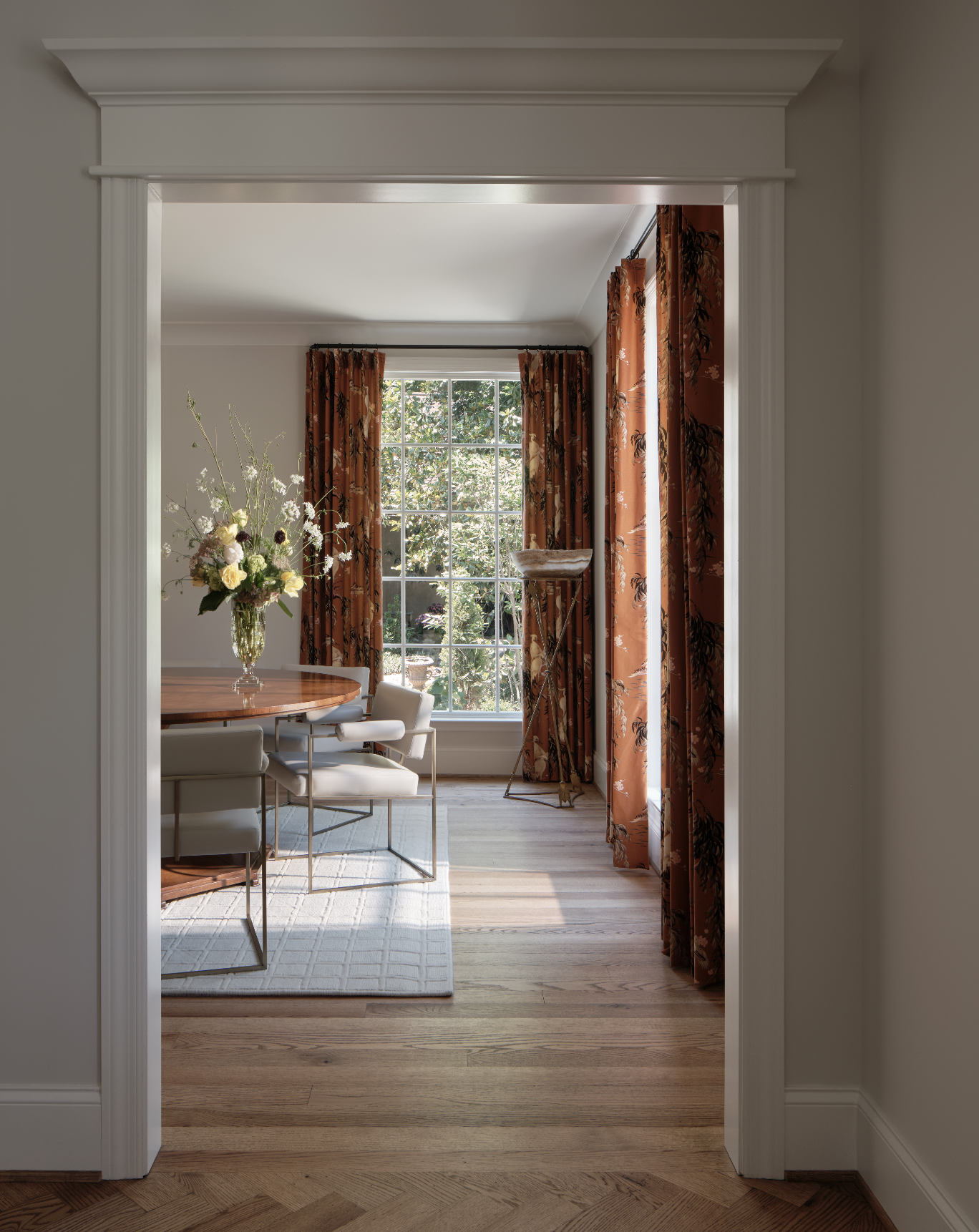 A dining room with wooden flooring, white walls, and large windows with orange floral curtains. There is a round wooden table with a vase of flowers, and white chairs. A floor lamp stands near the window.