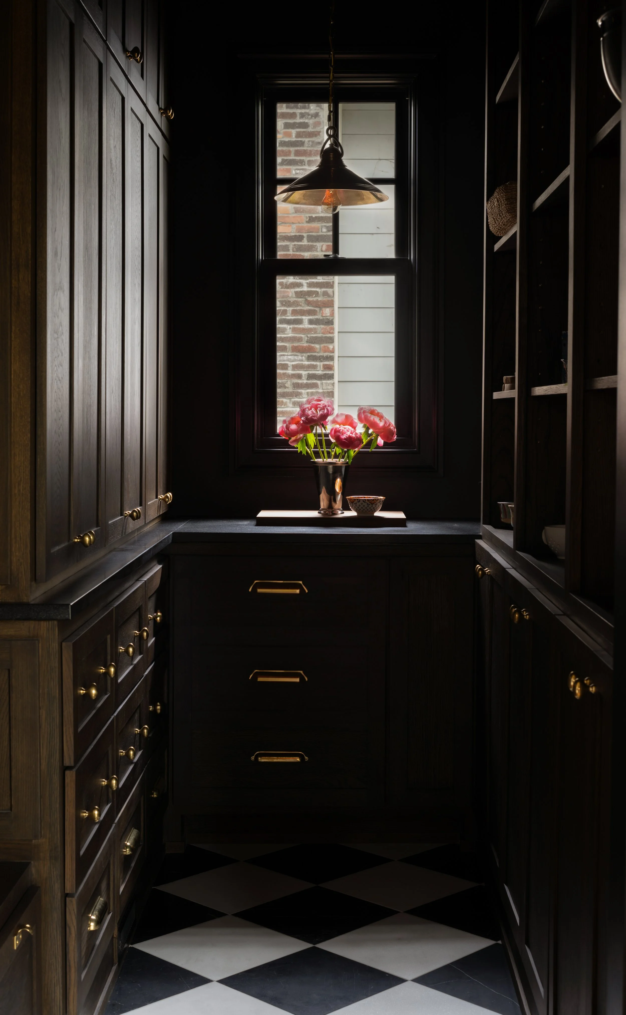 A narrow kitchen with dark wooden cabinets and a black and white checkered floor. A window is centered at the end, with a flower vase and pink flowers on the windowsill. A hanging light fixture is above.