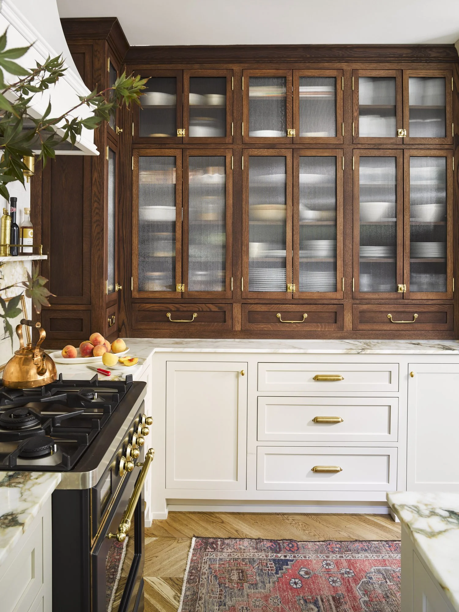 Kitchen with white lower cabinets, brass handles, a marble countertop, and wooden upper cabinets with glass doors showing dishes inside. A stove with brass accents, a copper teapot, and a bowl of peaches on the countertop.