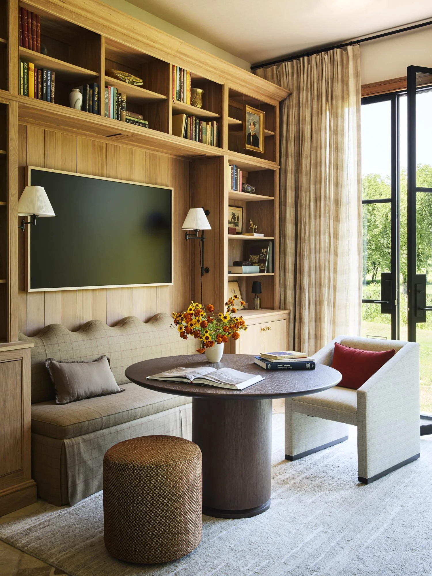 Living room with wooden built-in bookshelves, a large flat-screen TV, a beige sofa with beige and brown pillows, a round wooden table with a book and a flower vase, a white armchair with a red pillow, a small side table with a black lamp, and large w