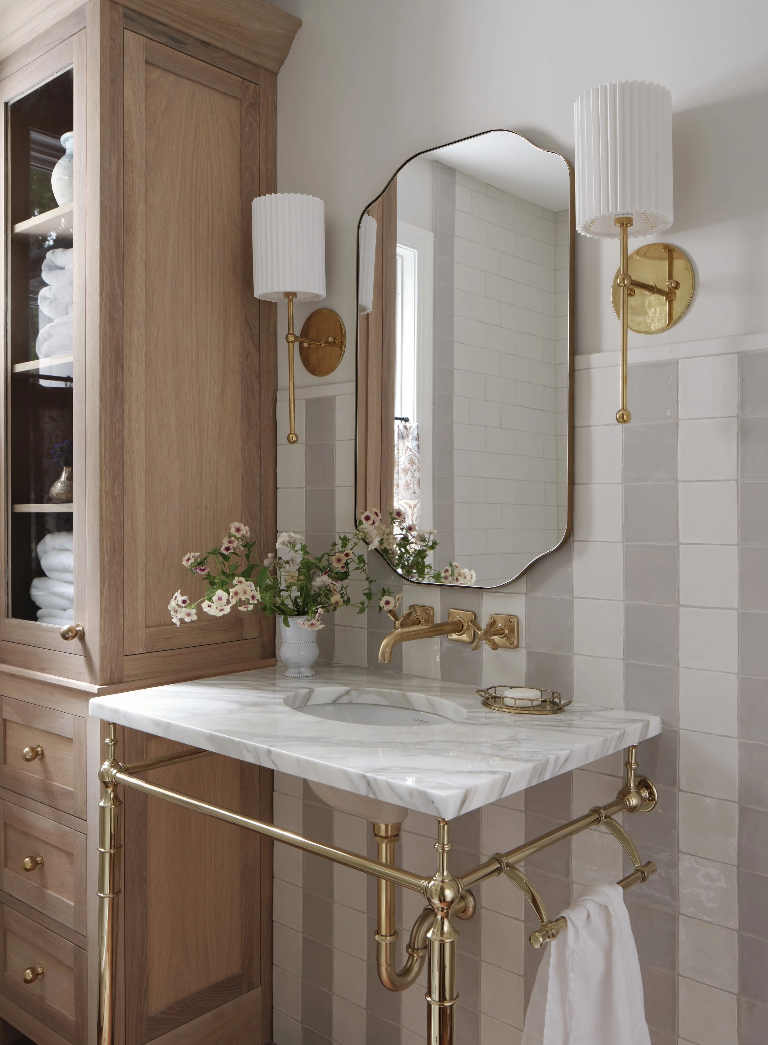 A bathroom with a marble sink, gold fixtures, a wooden cabinet, a mirror, and two wall-mounted gold and white light fixtures. There is a vase with flowers on the sink.