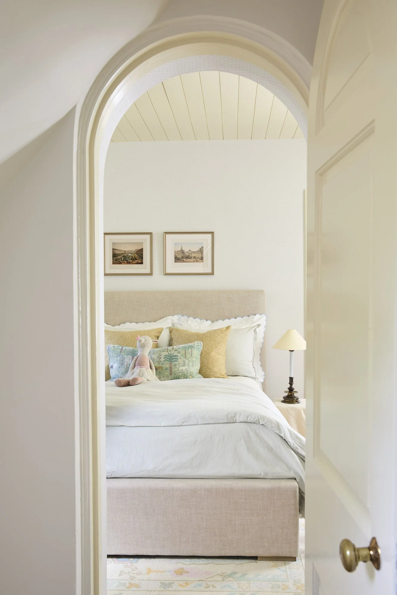 View through an open door into a well-decorated bedroom with a cream-colored bed, patterned throw pillows, a stuffed llama, two framed pictures on the wall, a table lamp, and a soft rug on the floor.