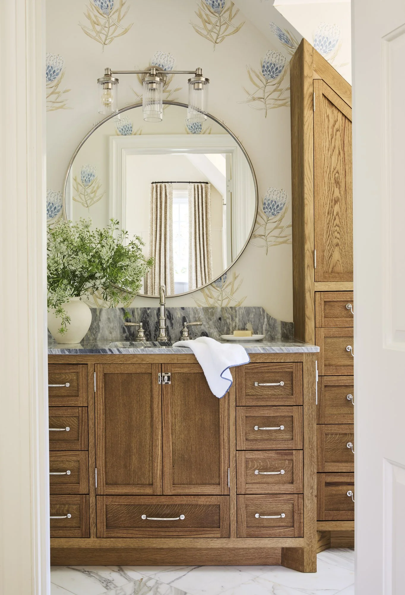 A bathroom vanity with a marble countertop, a round mirror, a vase of green plants, and a towel. Wooden cabinetry and a patterned wallpaper with blue and beige flowers. A window with curtains is reflected in the mirror.