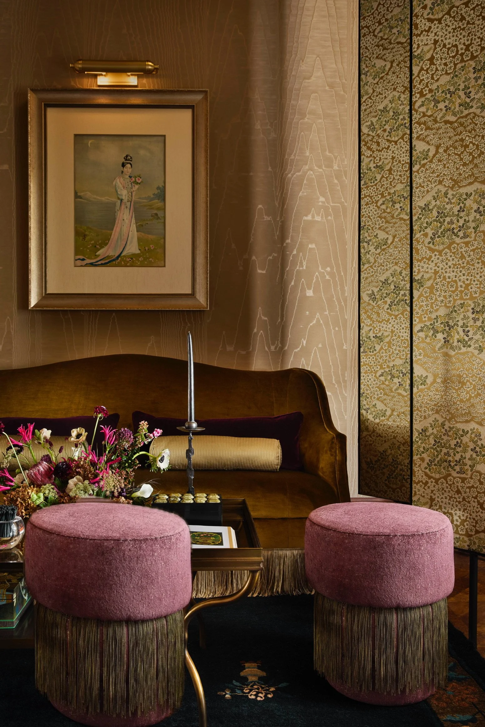 Cozy living room corner with two pink upholstered stools, a dark brown vintage sofa with a yellow pillow and a dark purple pillow, a side table with a floral arrangement and a candle holder, a framed painting of a woman in traditional dress on the wa