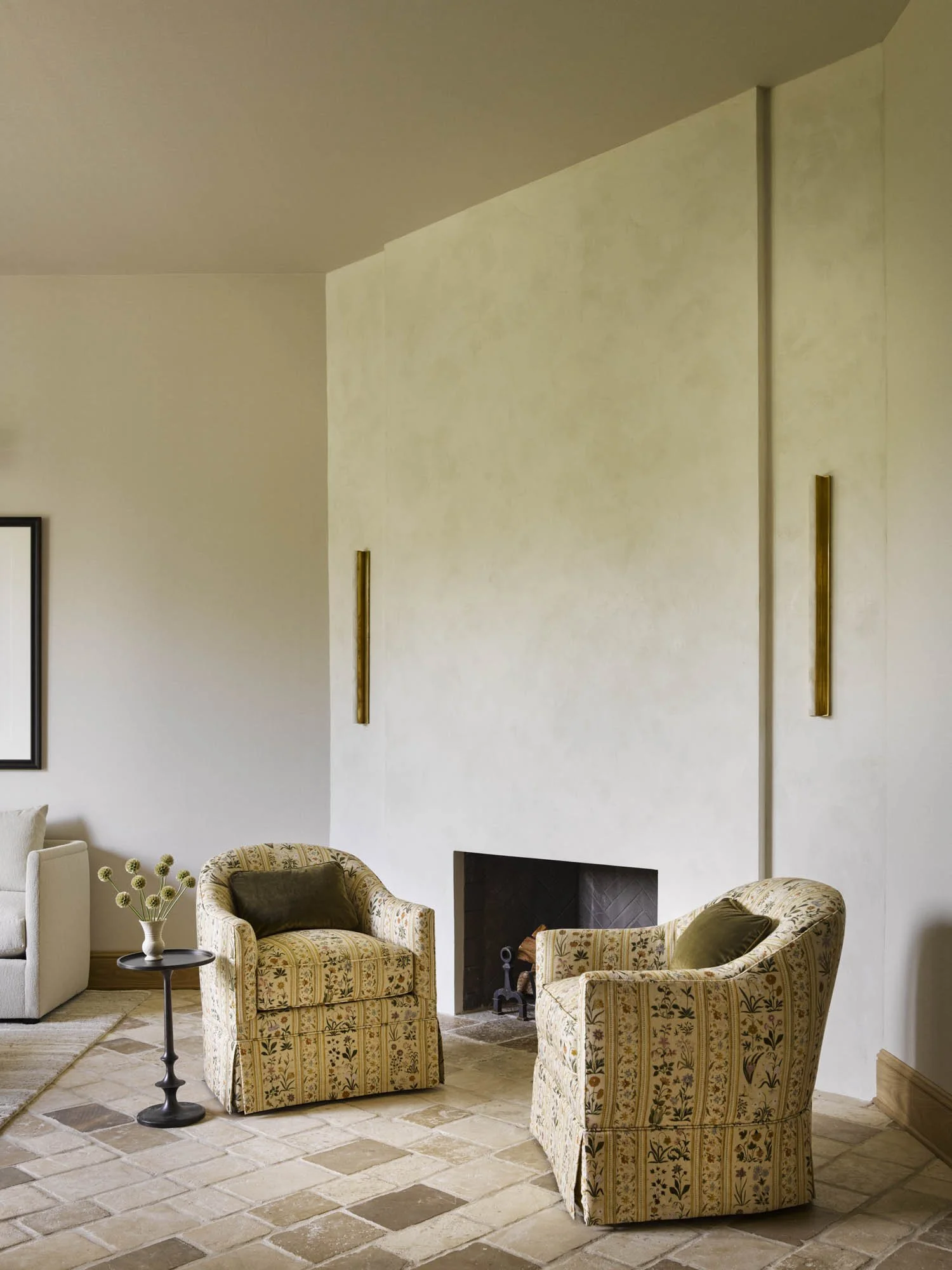 Living room with two patterned armchairs, a small table with a vase of flowers, a fireplace, and partial view of a sofa.