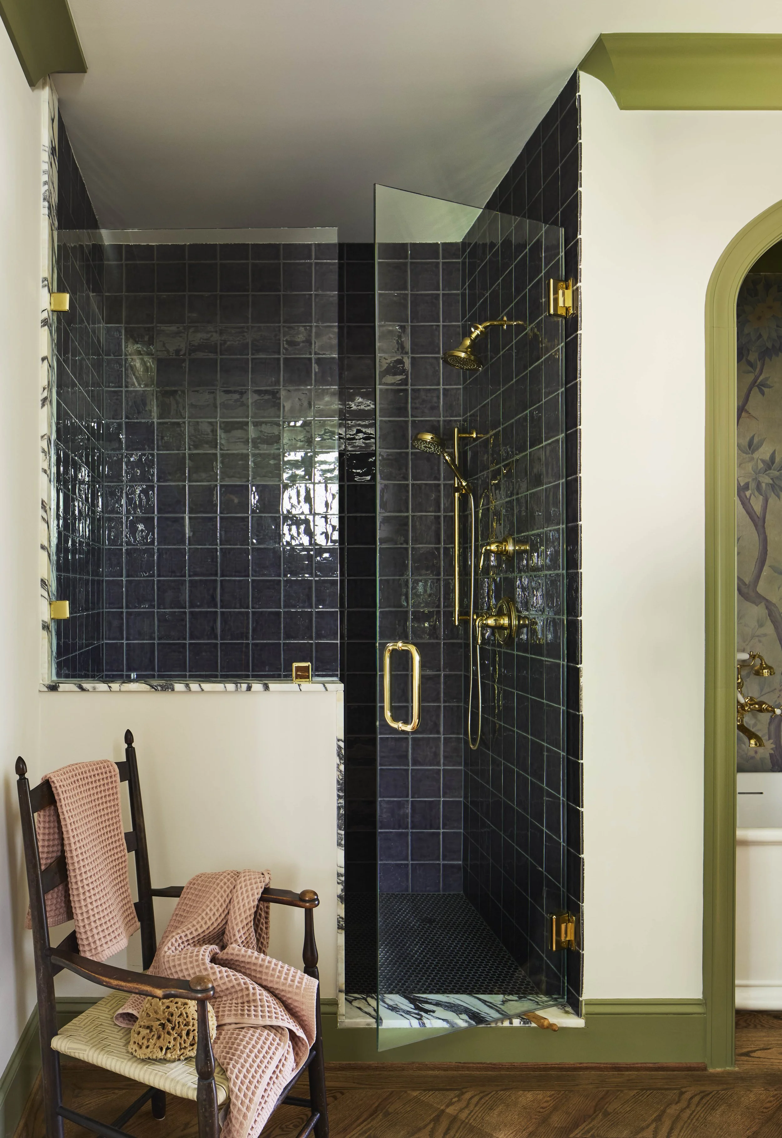 A glass-enclosed shower with black tiles, gold fixtures, and two shower heads. There is a wooden chair with pink towels draped over it in front of the shower, on a wooden floor.