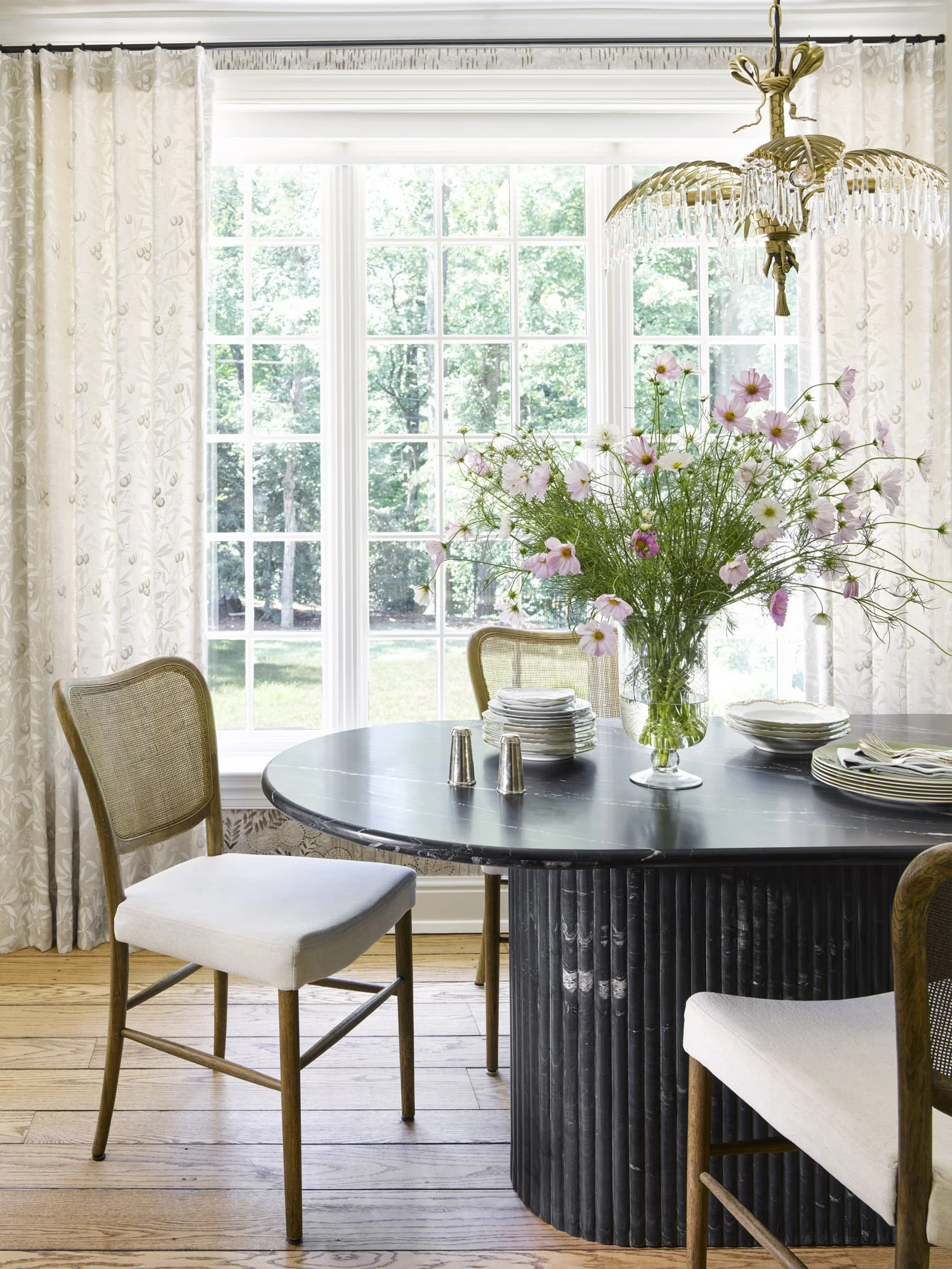 A dining room with a round black marble table, surrounded by four cream-colored chairs, a vase with pink and white flowers, plates, silverware, salt and pepper shakers, large windows with white trim, and cream floral curtains. There's a chandelier ab
