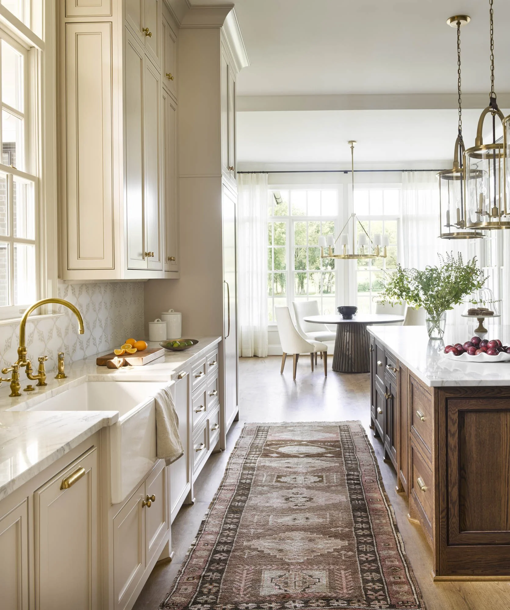 Bright kitchen with white cabinets, brass fixtures, and a rustic wooden island, connecting to a dining area with large windows and light curtains.