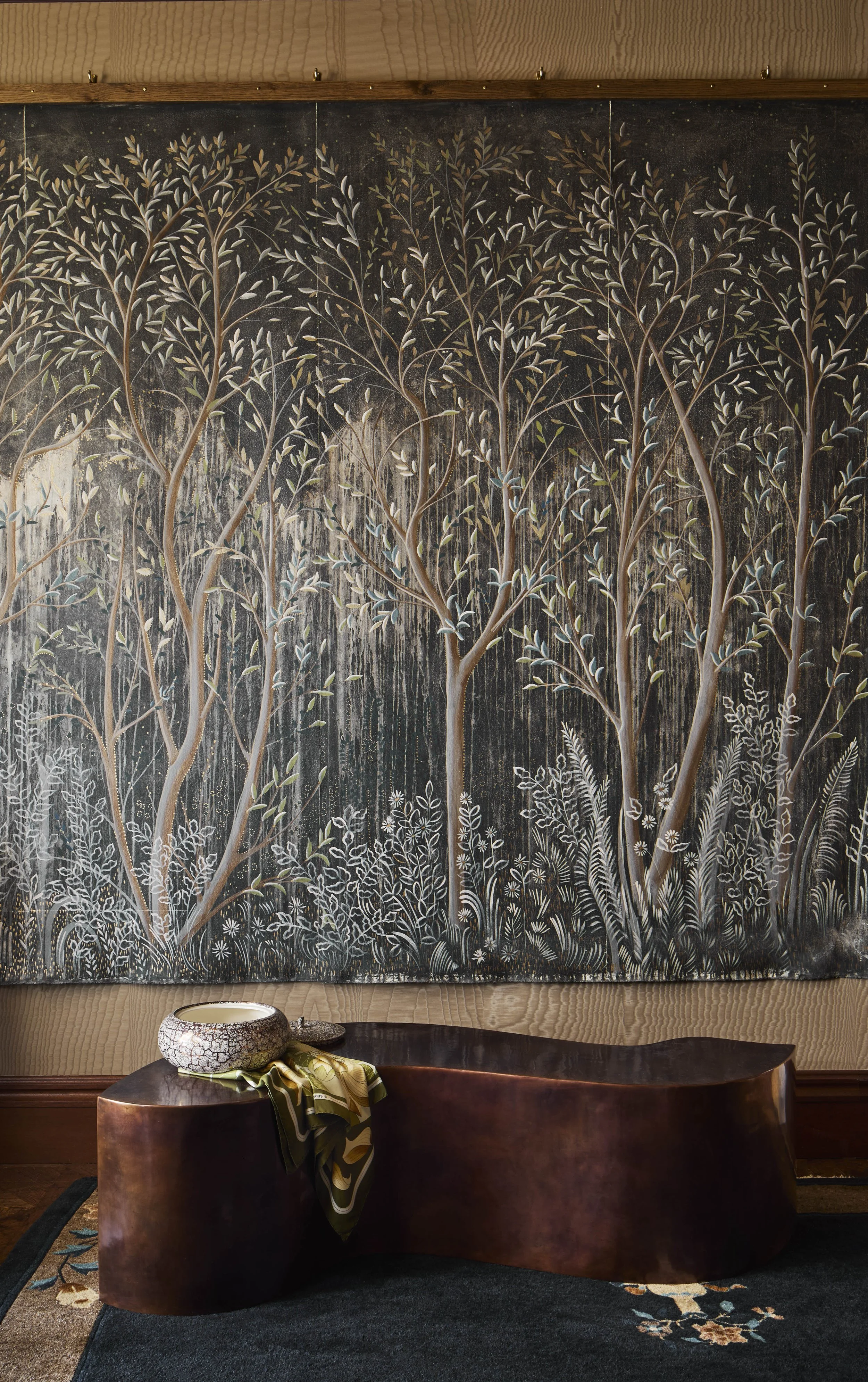 A dark painting of tall trees with slender trunks and leaves, set behind a leather-upholstered bench and a decorative bowl with a scarf on top in a warmly lit room.