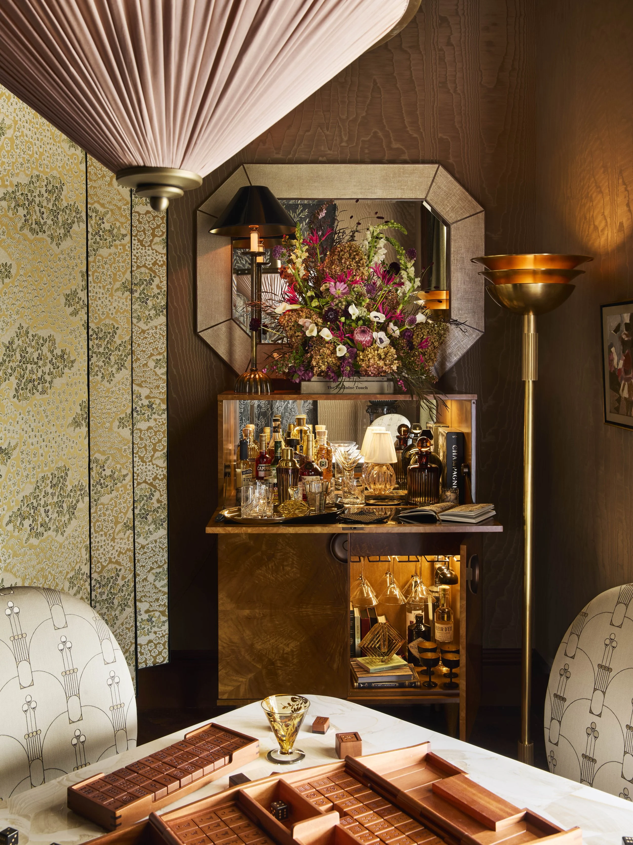 A luxurious game room with a hexagonal mirror, filled with flowers and a bar setup, surrounded by elegant chairs and a table with chocolate tiles and a glass with a gold design.