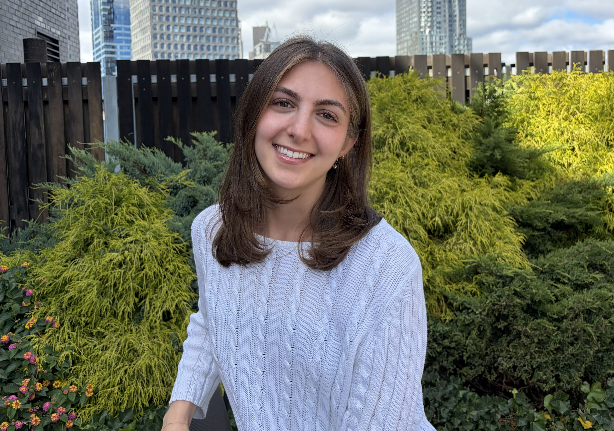 Smiling woman in a white sweater posing in a rooftop garden with city skyscrapers in the background
