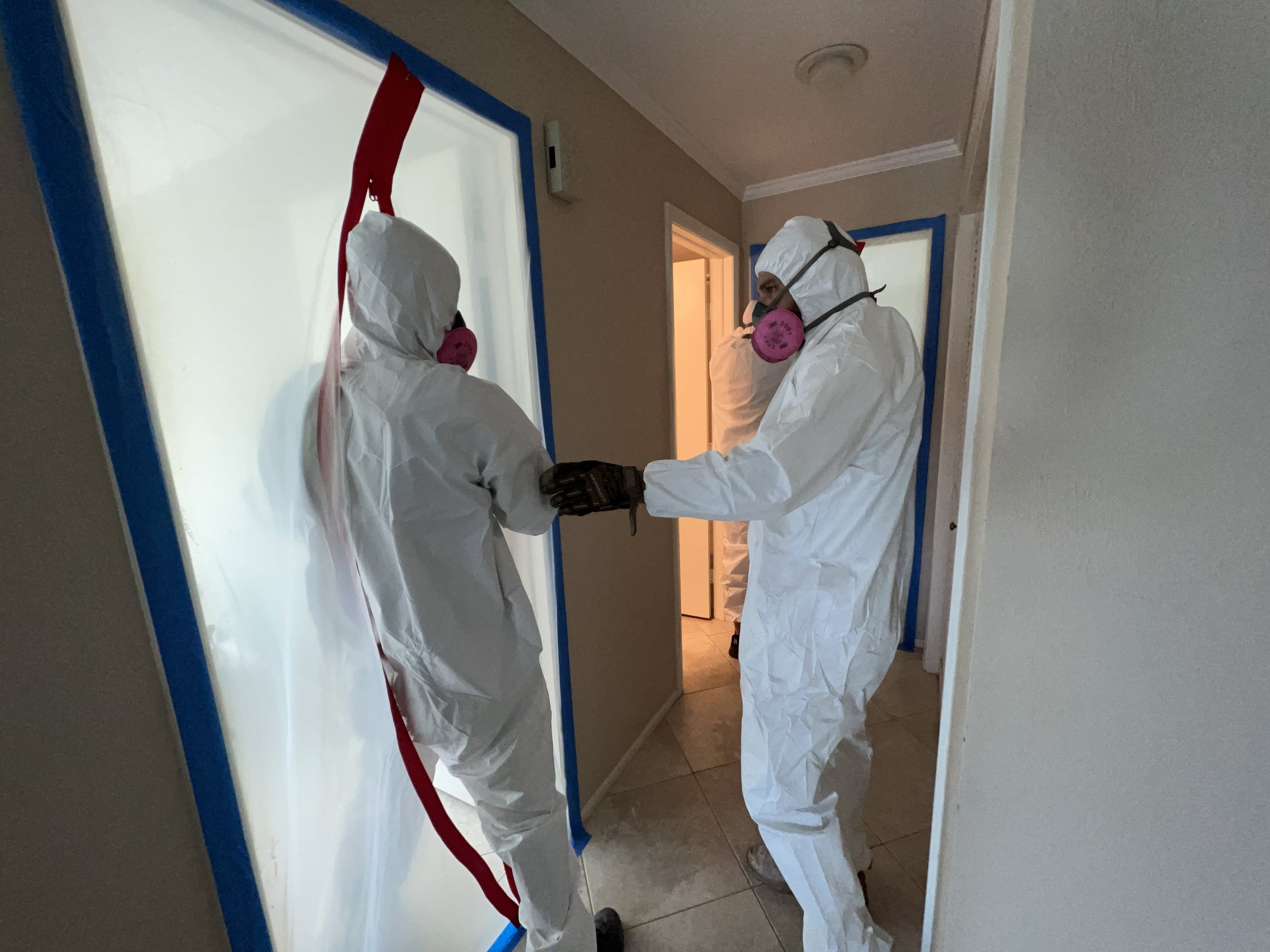 Two individuals in full personal protective equipment (PPE), including white hazmat suits and pink respirators, are working together in a room with beige walls and tiled flooring. They are behind a blue and white barrier with a red hose attached, possibly in a healthcare or decontamination setting.
