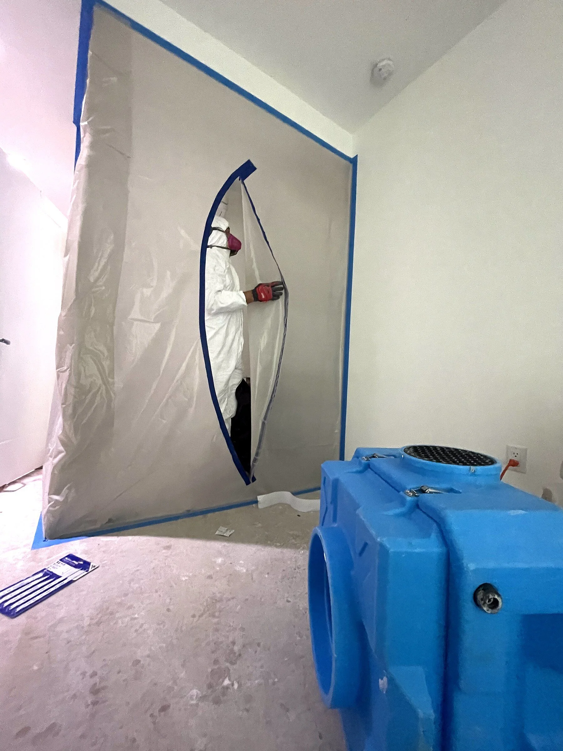 A worker in protective gear painting a wall in a room, with a blue plastic trash bin in the foreground and painter's tape outlining the wall.