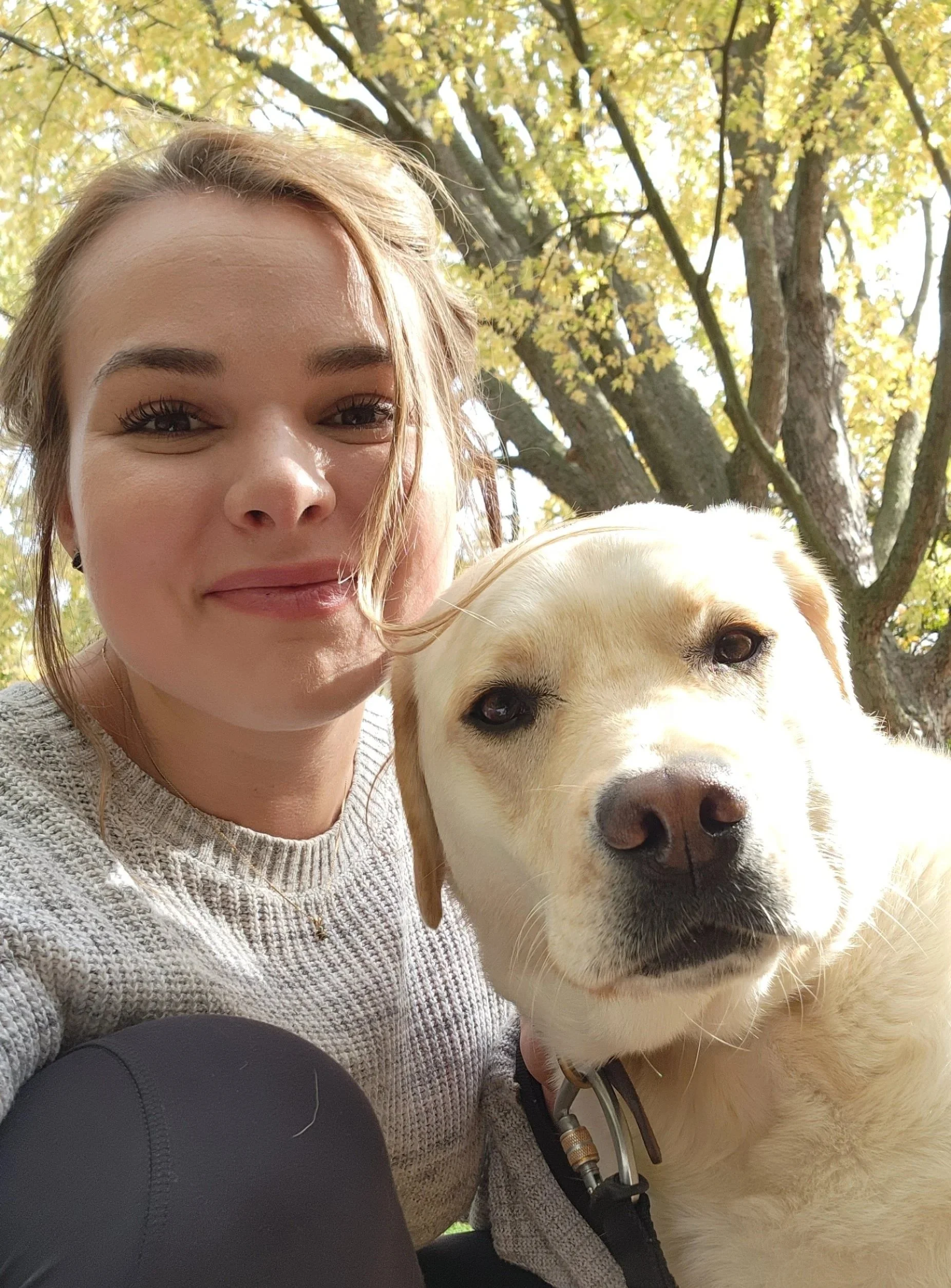 A woman with light brown hair smiling, taking a selfie with a Labrador retriever dog outdoors near a large tree with yellow leaves.