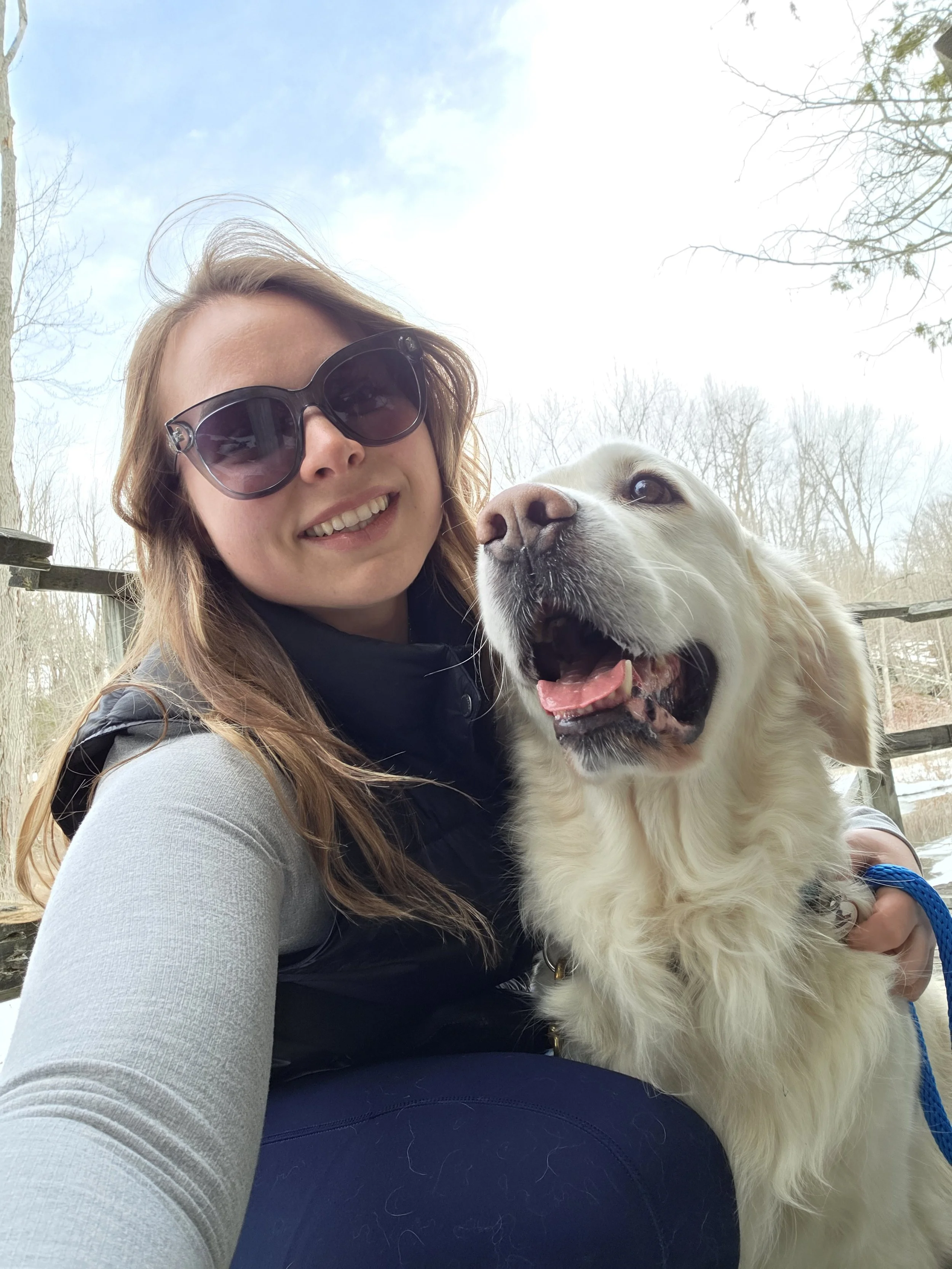 A woman with long blonde hair, dark sunglasses, and a big smile taking a selfie with a large white retriever dog outdoors, with leafless trees and a bright sky in the background.