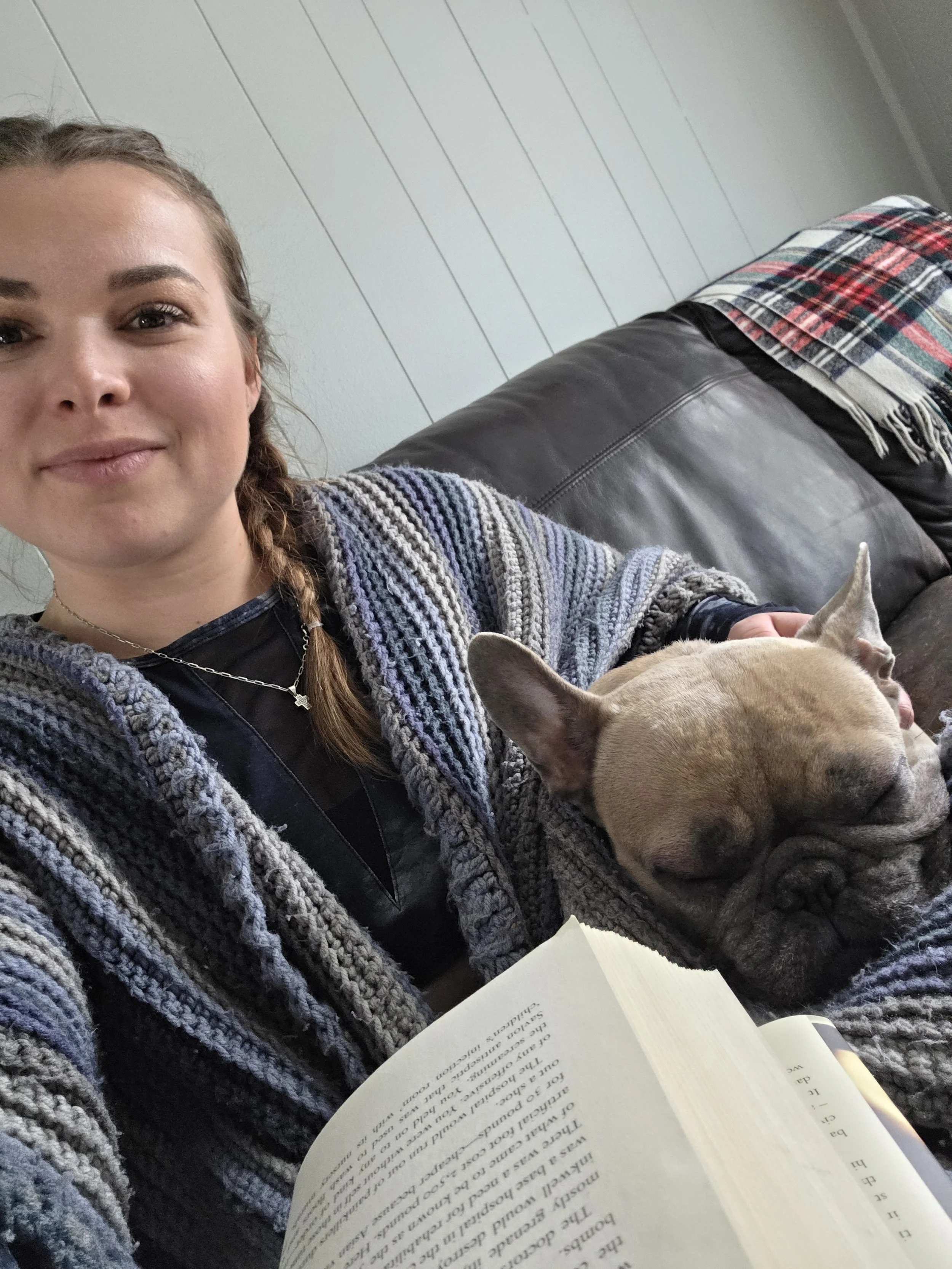 A woman with braided hair wearing a striped gray sweater and a silver necklace is sitting on a couch, reading a book. A tan-colored dog is resting on her arm with its eyes closed. There is a colorful plaid blanket on the back of the couch and white wood-paneled wall in the background.
