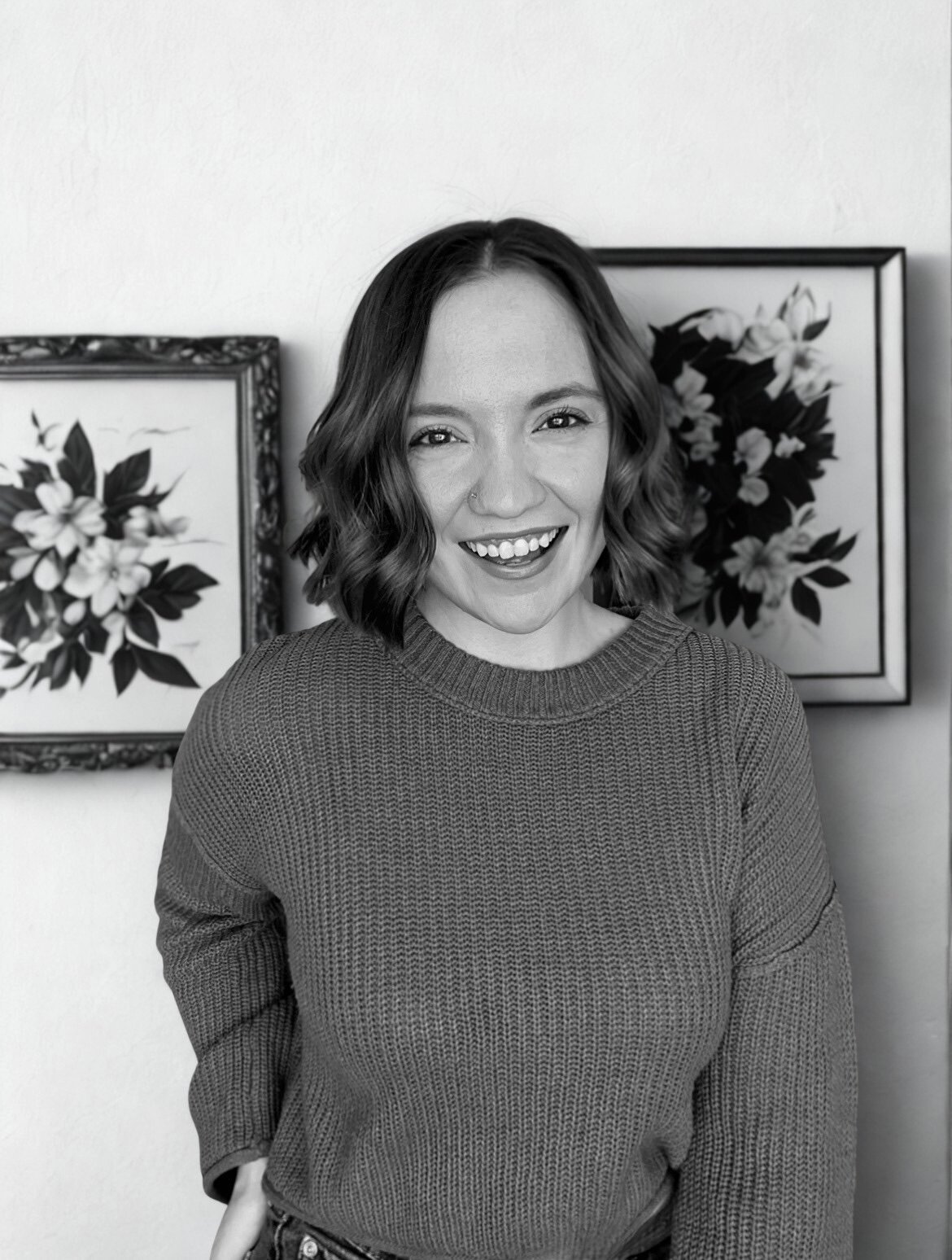 A woman with shoulder-length wavy hair smiling and wearing a sweater, standing in front of two framed floral paintings.