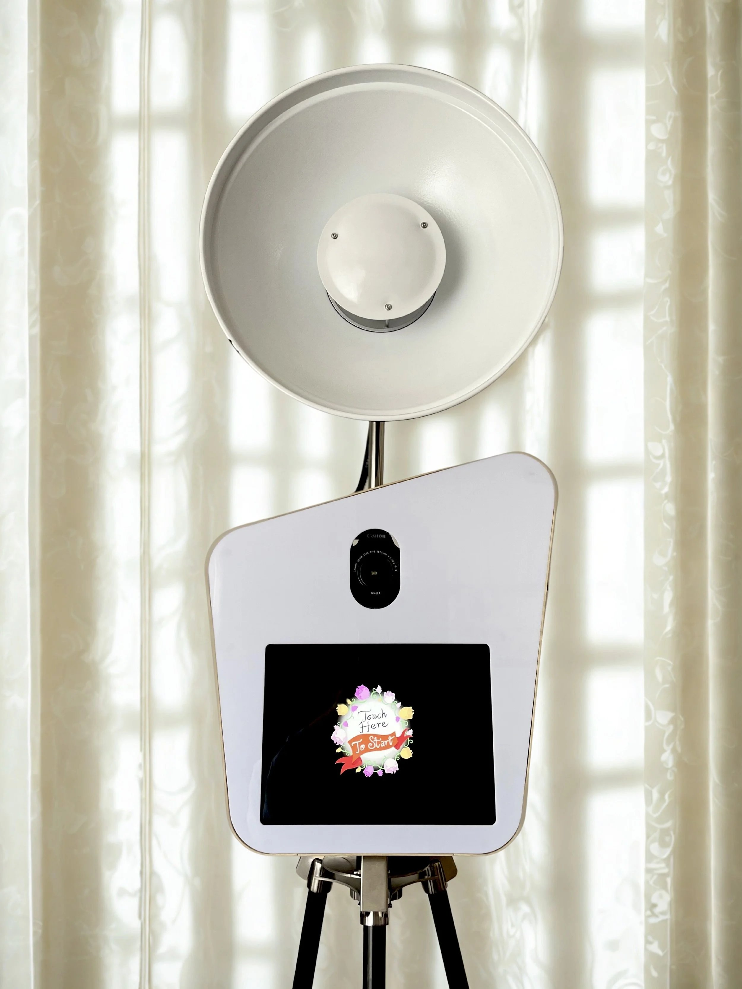 Photo of a white photo booth with a circular light on top, a camera in the middle, and a touchscreen at the bottom displaying a colorful display that says 'Touch Here To Start', with curtains in the background.