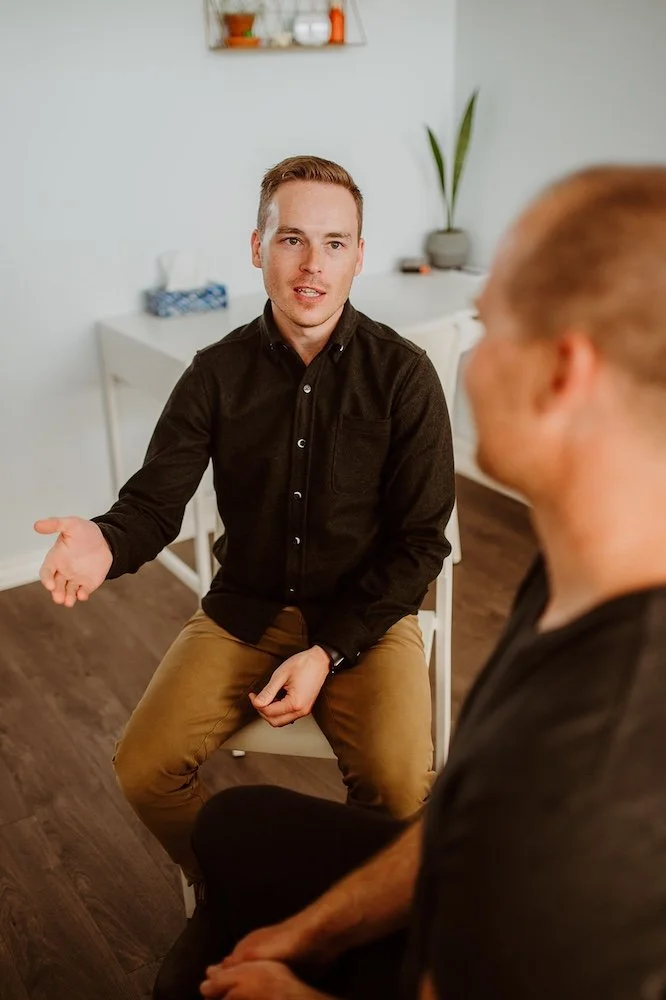 Two men have a conversation in a room, with one gesturing with his hand and the other listening. The room has a white cabinet, a potted plant, and some decorative items in the background.