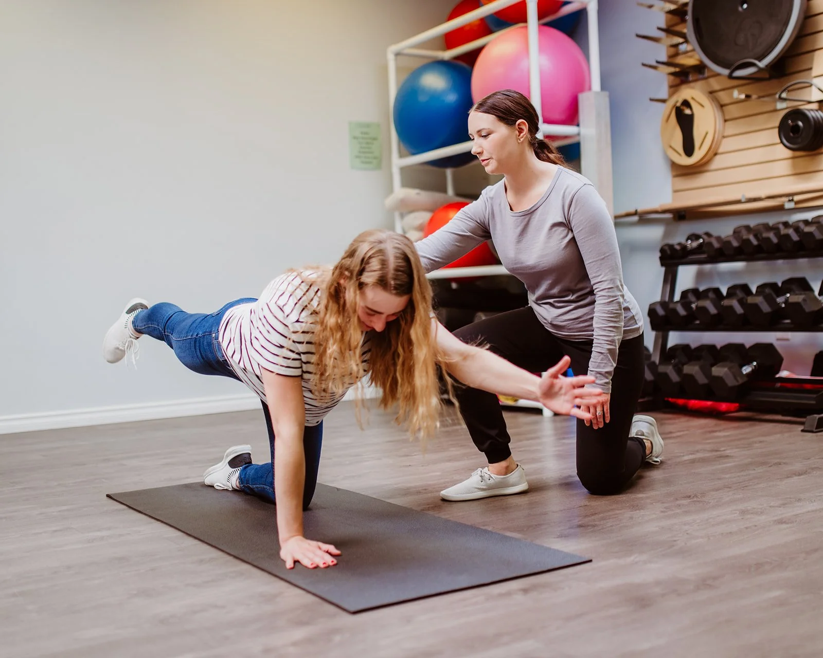 A woman assists a girl with a fitness exercise on a yoga mat in a gym. The girl is in a plank position, extending one arm forward and the other supporting her weight. The woman is supporting the girl, guiding her back and providing stability.