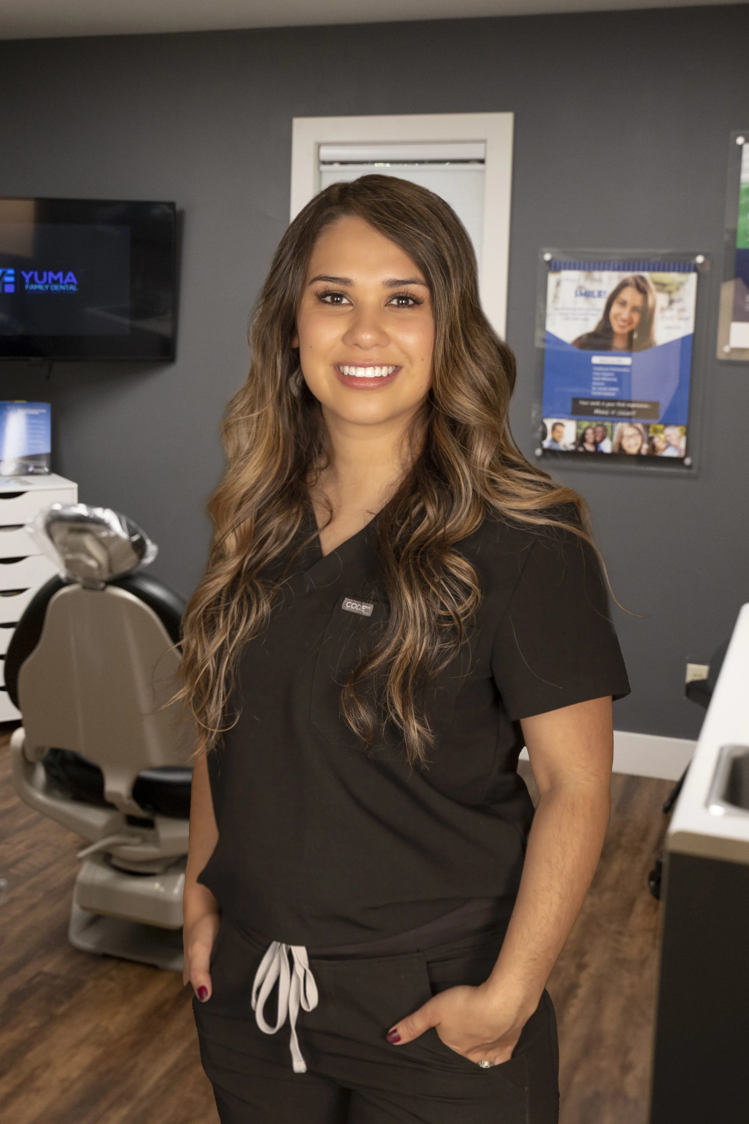 A smiling woman in black scrubs standing in a dental office with dental chair and posters in the background.