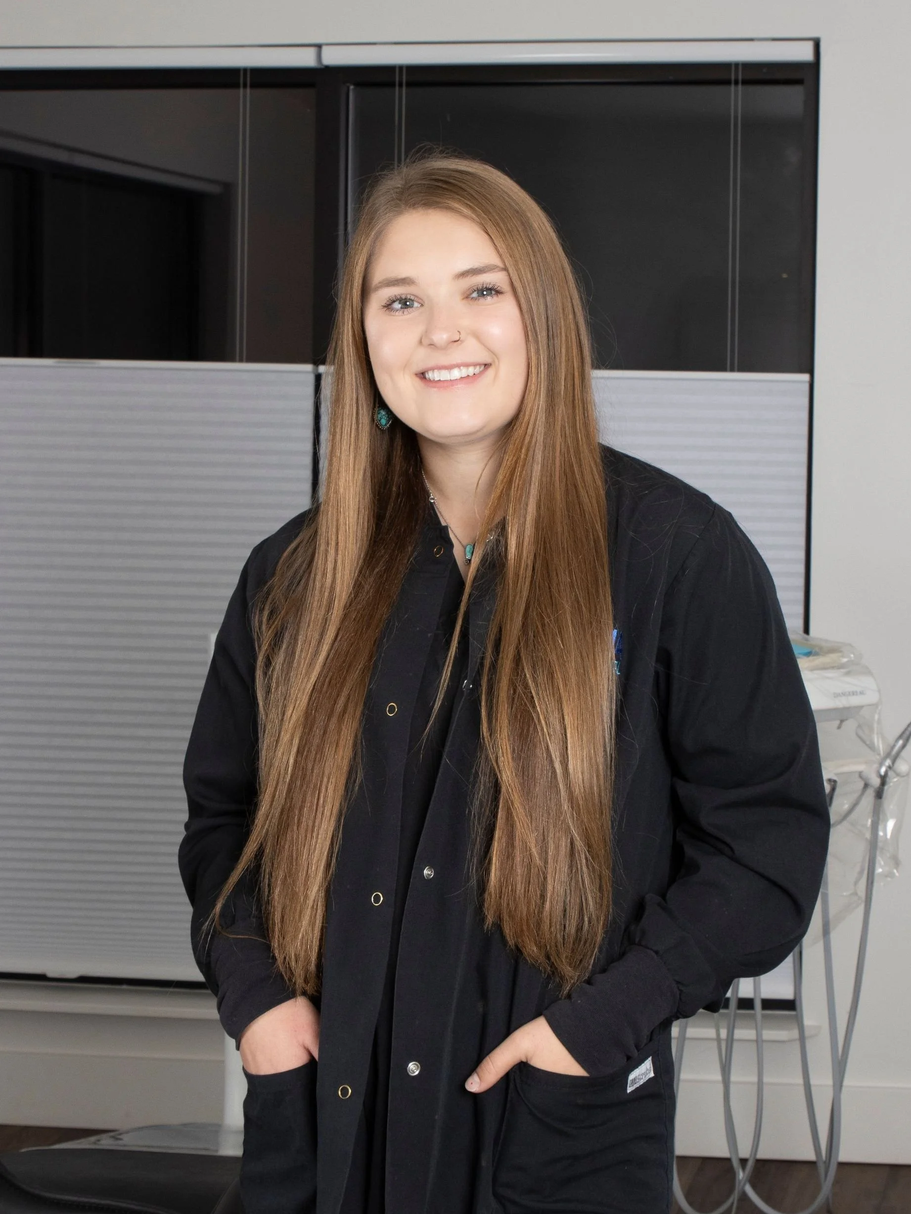 A young woman with long brown hair, wearing a black jacket, smiling and standing with her hands in her pockets in an indoor setting.