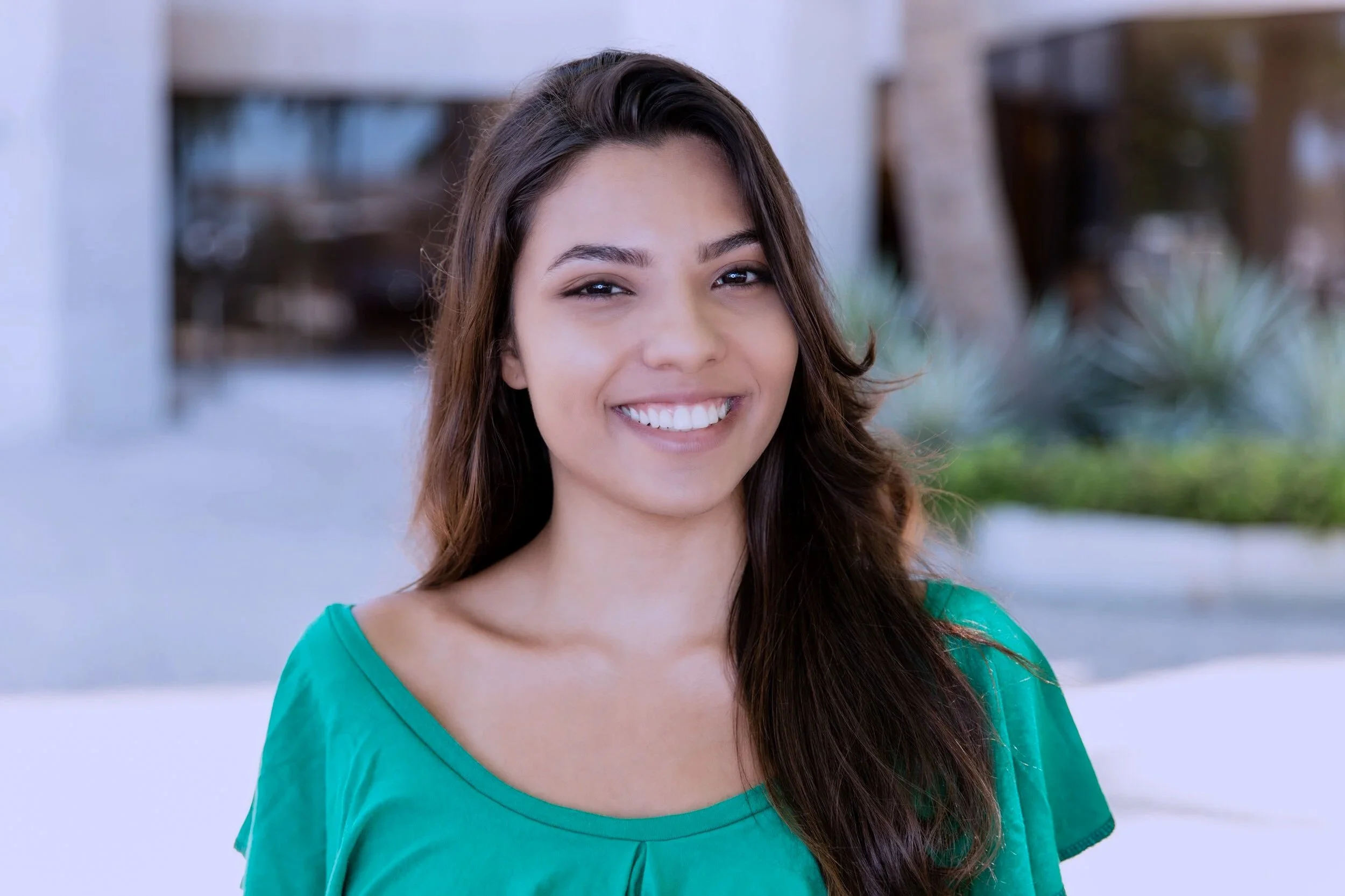 A woman with long brown hair smiling outdoors, wearing a green top with a background of a building and greenery.