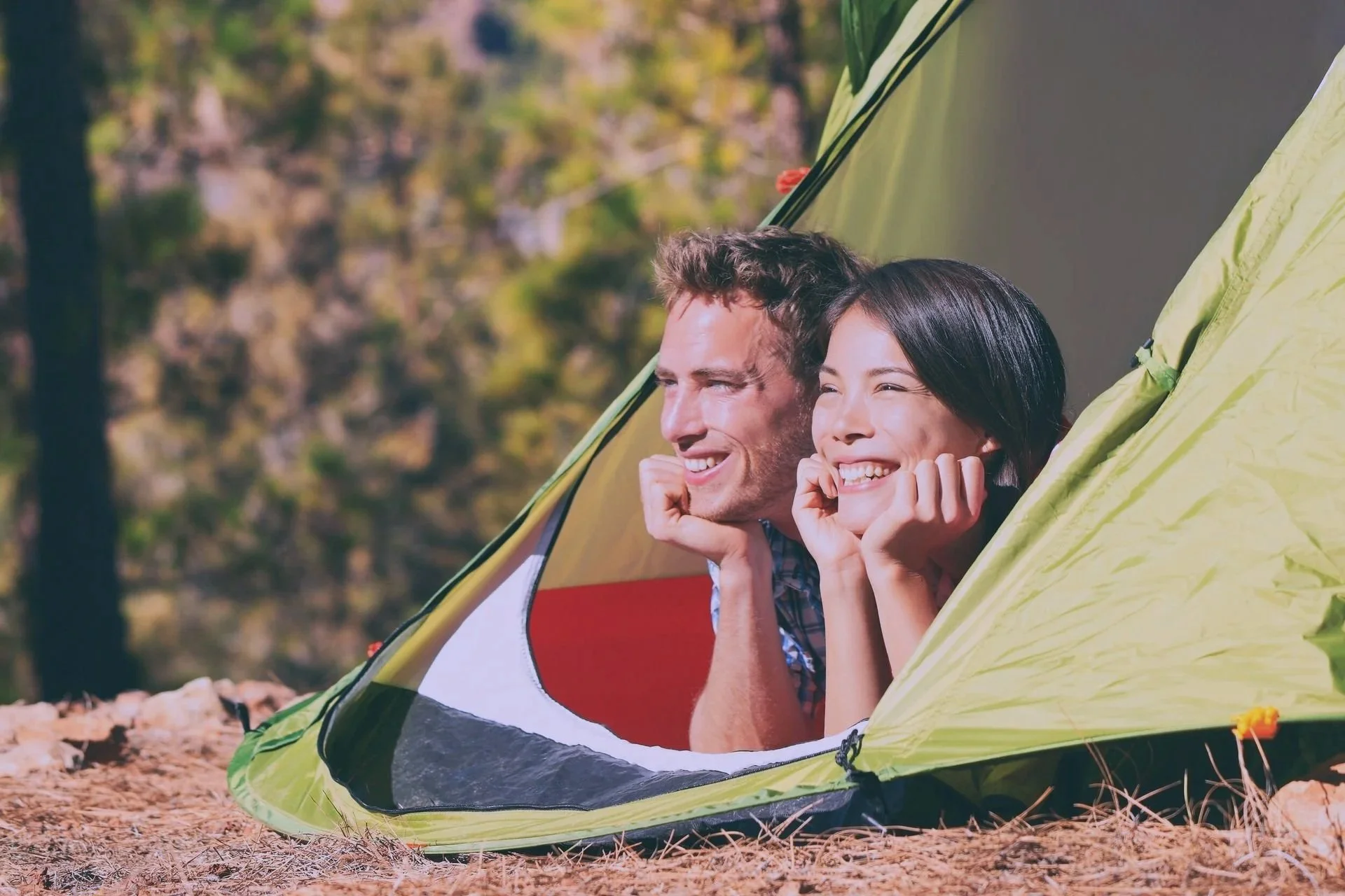 Two smiling people lying inside a tent in an outdoor forest setting.