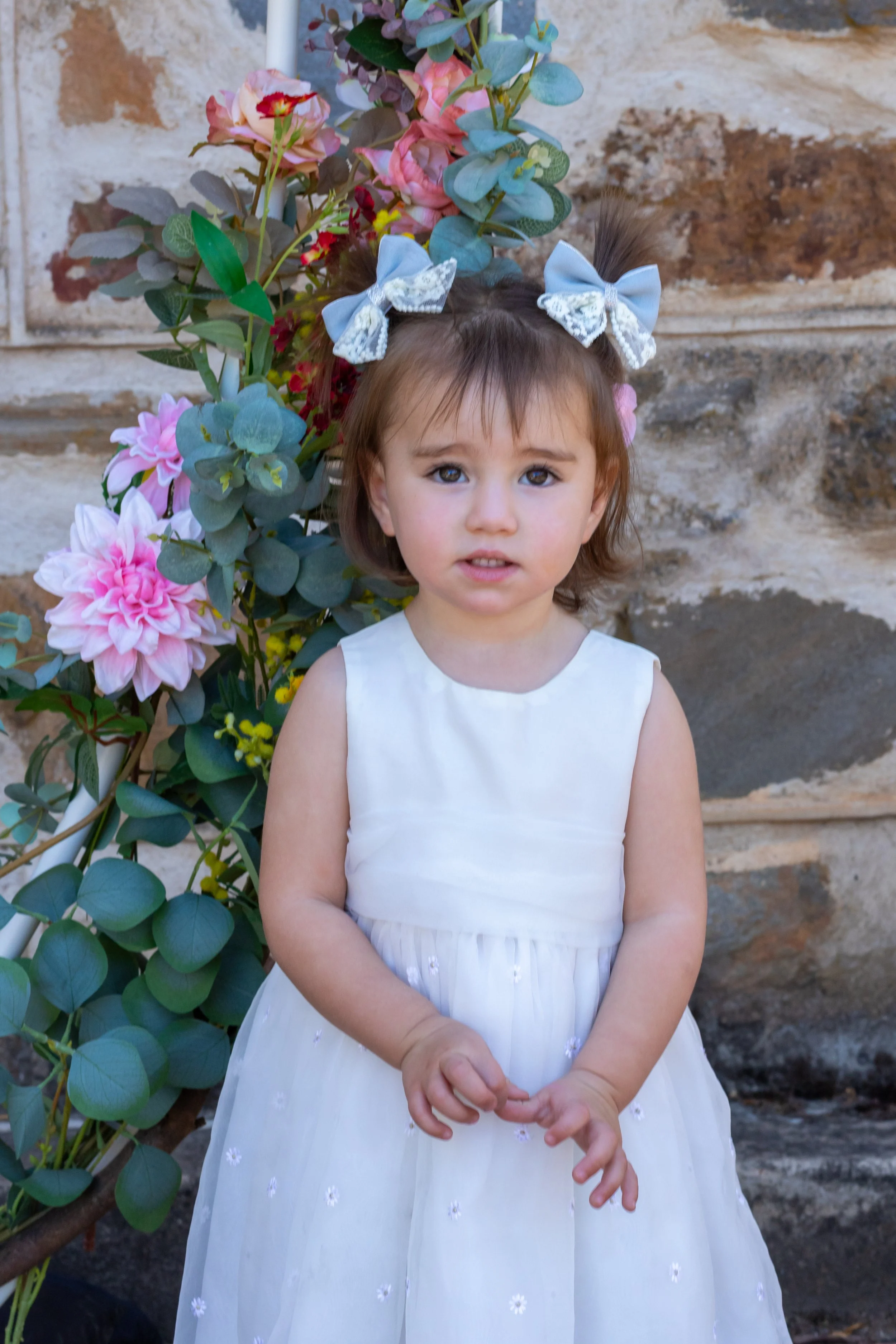 A young girl wearing a white dress with small floral details, standing in front of a floral arrangement with pink and purple flowers and green foliage, against a stone wall background.
