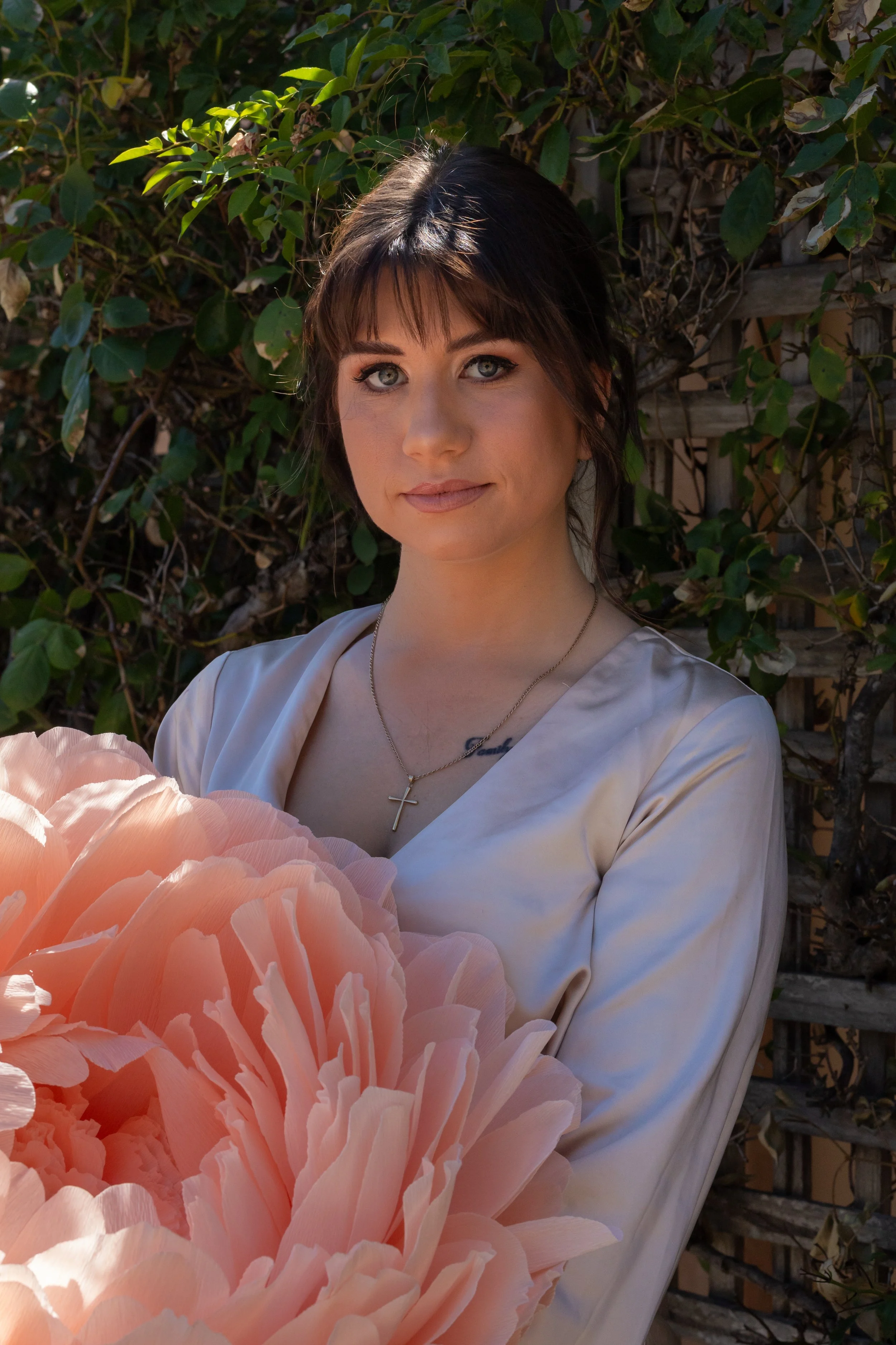 A woman with dark brown hair and blue eyes holding large pink peonies outdoors, with green leaves and a lattice in the background.