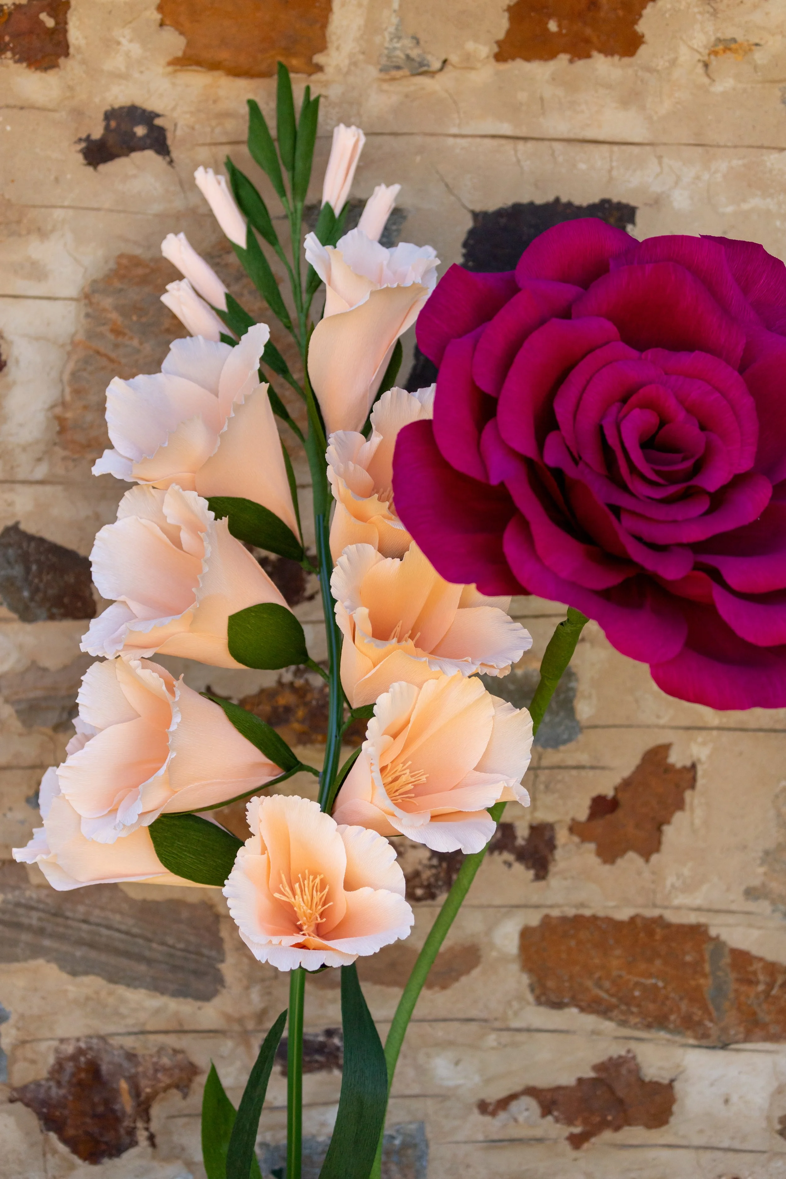 A close-up of a bouquet with peach-colored gladiolus flowers and a single deep pink rose against a rustic brick wall.
