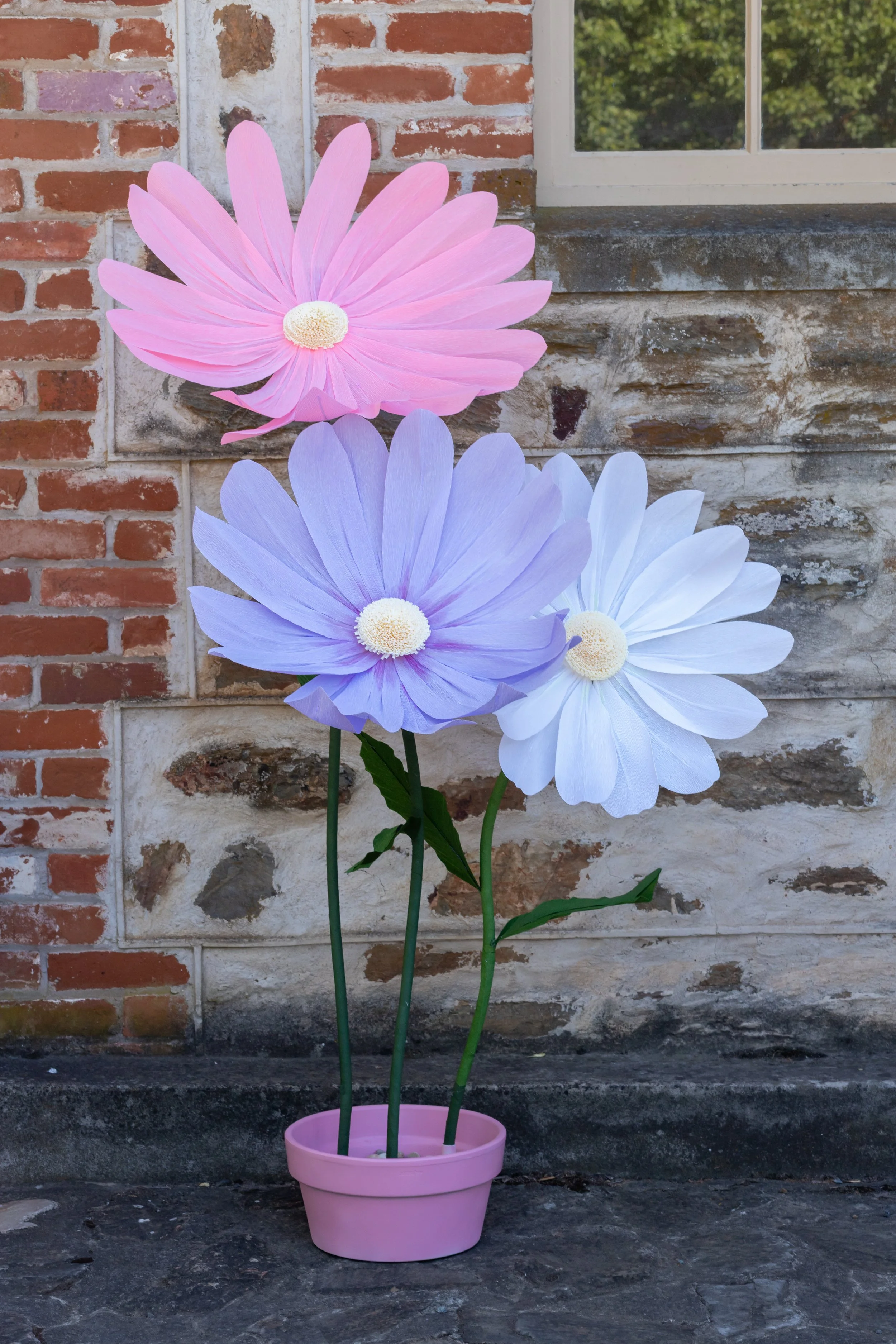 Artificial pink, purple, and white daisies in a pink pot placed on a stone surface against a brick and stone wall background with a window.