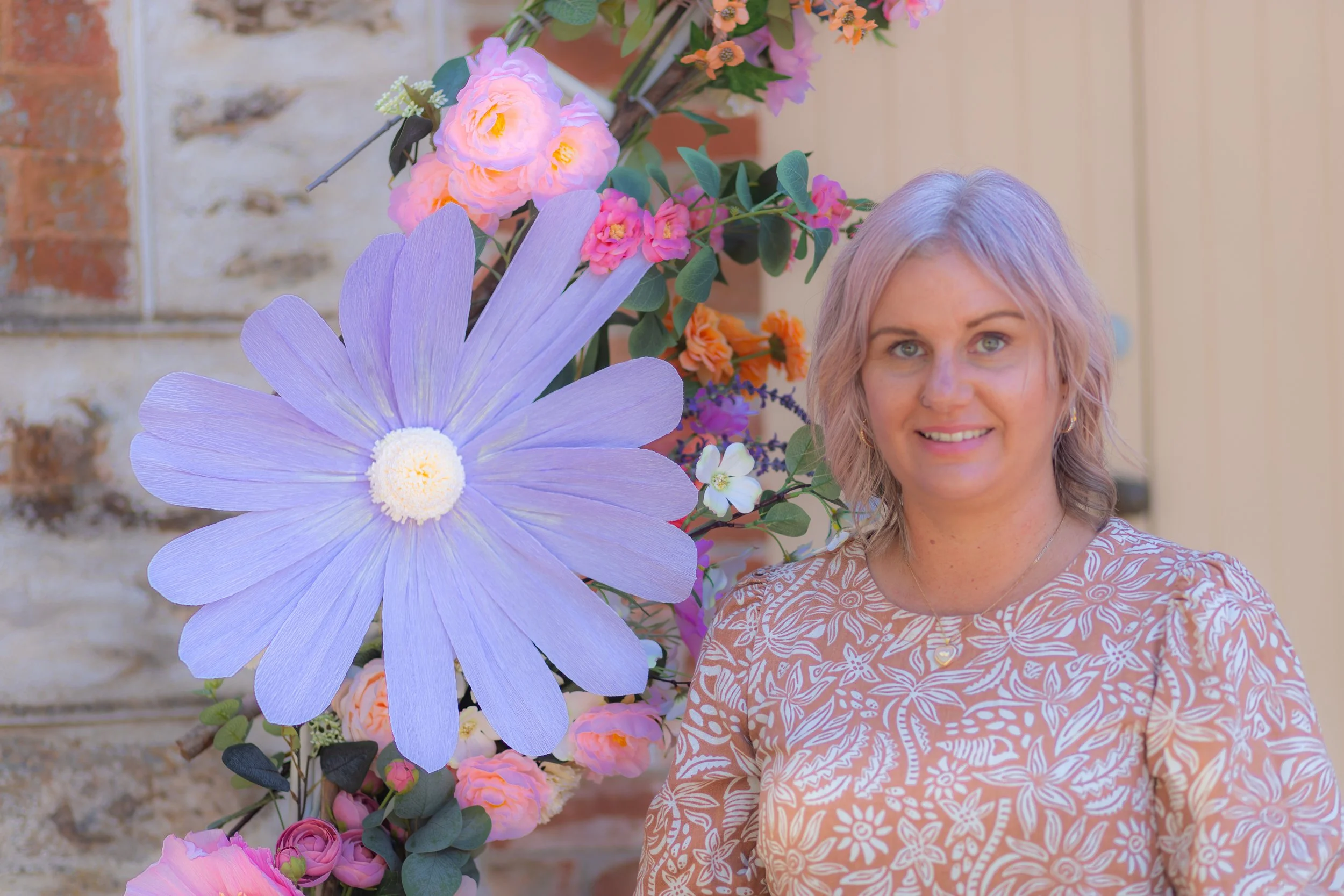 A woman with short, wavy, light-colored hair and earrings, smiling, standing next to a large arrangement of colorful flowers including a big purple flower, pink roses, and white blossoms, with a brick and wooden wall in the background.
